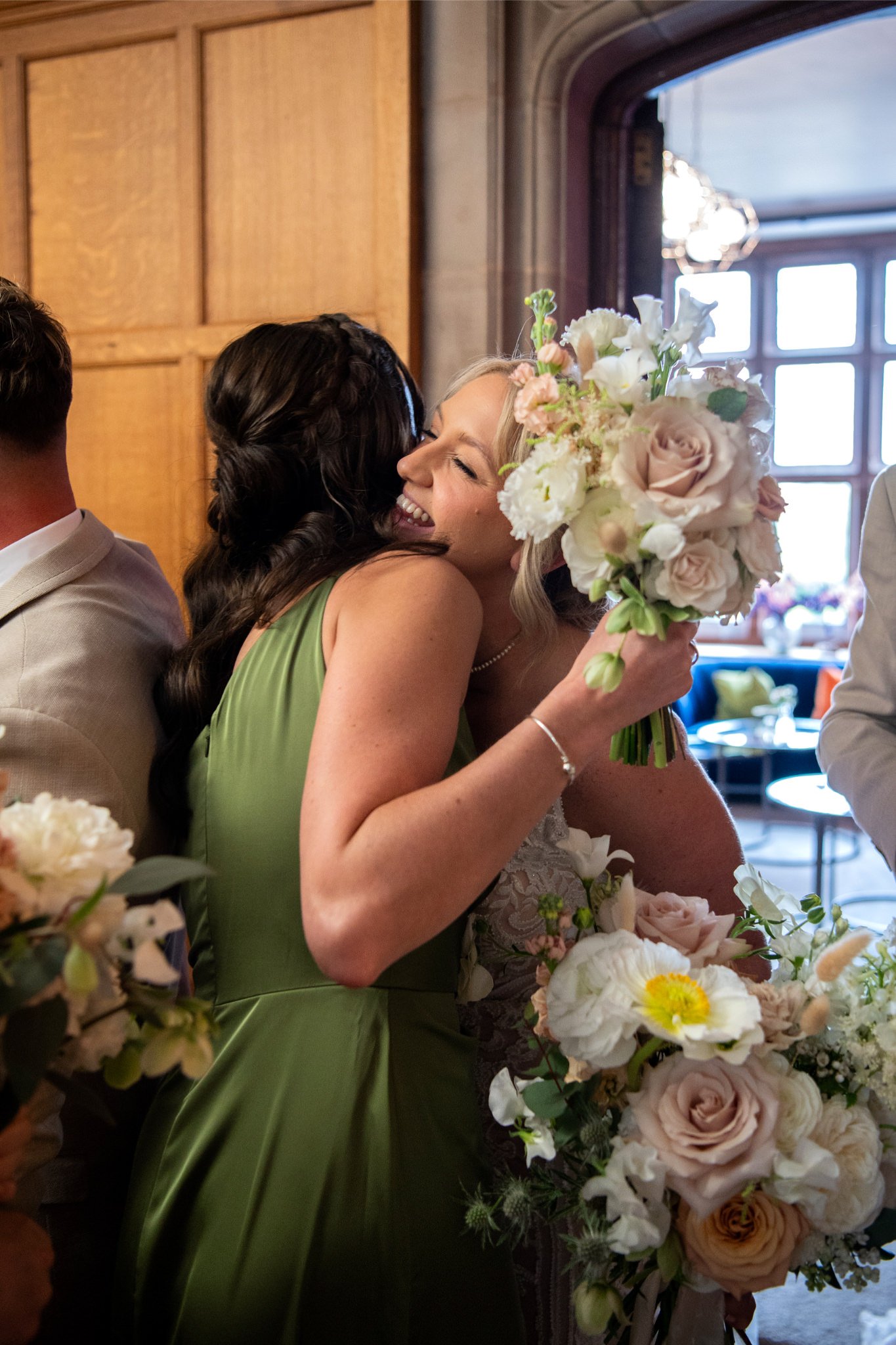 Two women hugging, one in a green dress and the other in a white dress, holding a bouquet of flowers, at a celebration or wedding in a warmly lit room.