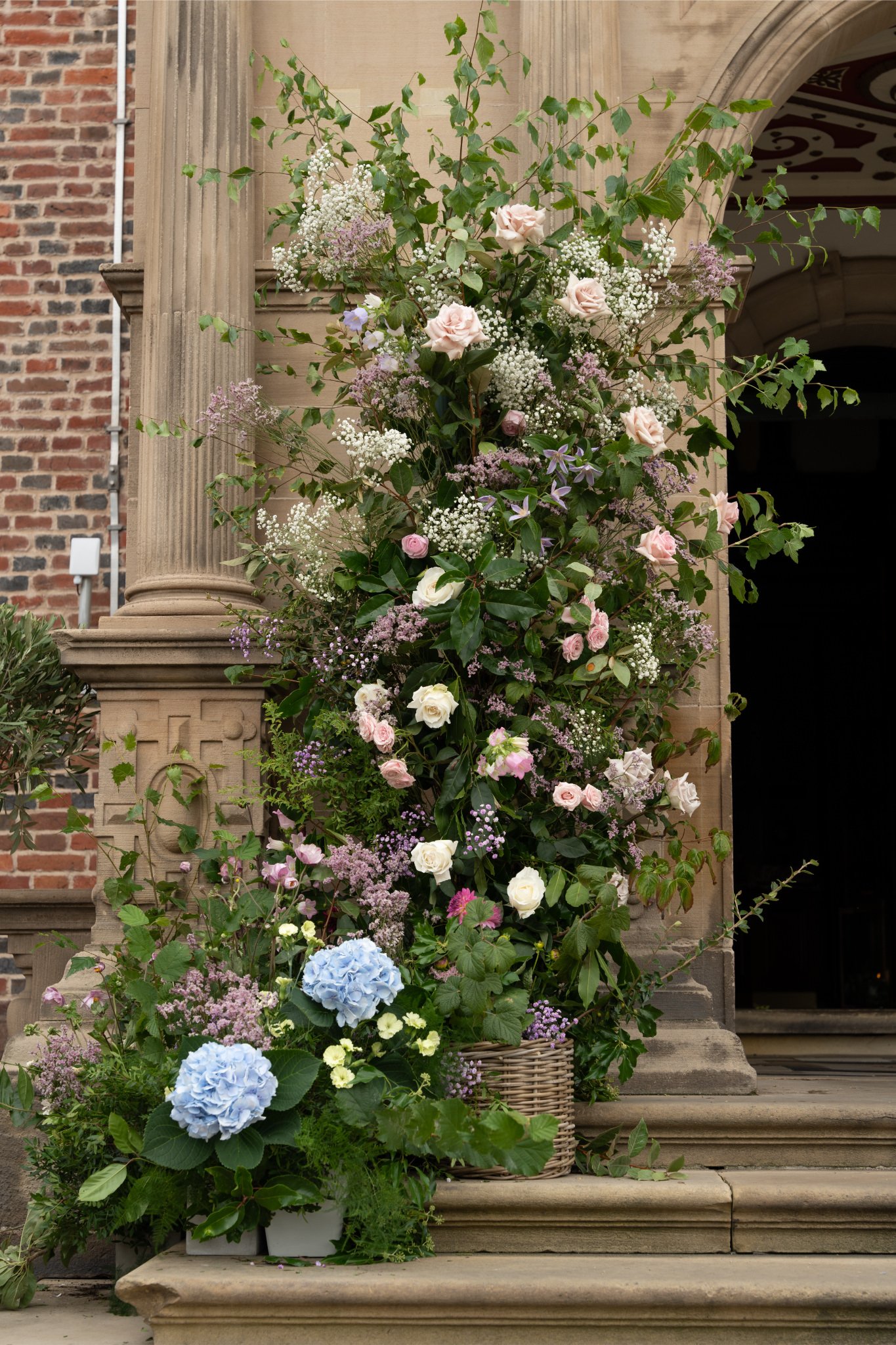 A large arrangement of pink and white roses, purple and white baby's breath, and blue hydrangeas on the steps of a building with stone and brick architecture.