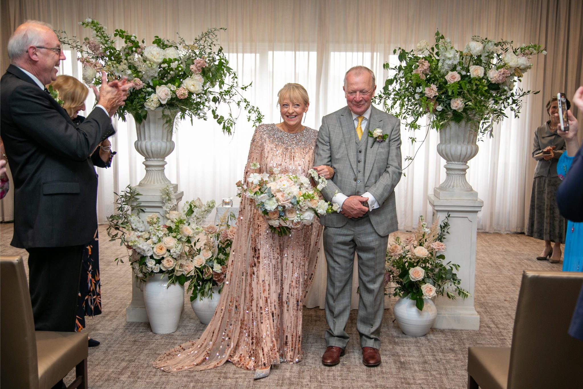 An elderly woman in a sparkling blush gown holding a large bouquet of flowers walking down the aisle with a man in a gray plaid suit, at a wedding ceremony decorated with large floral arrangements.