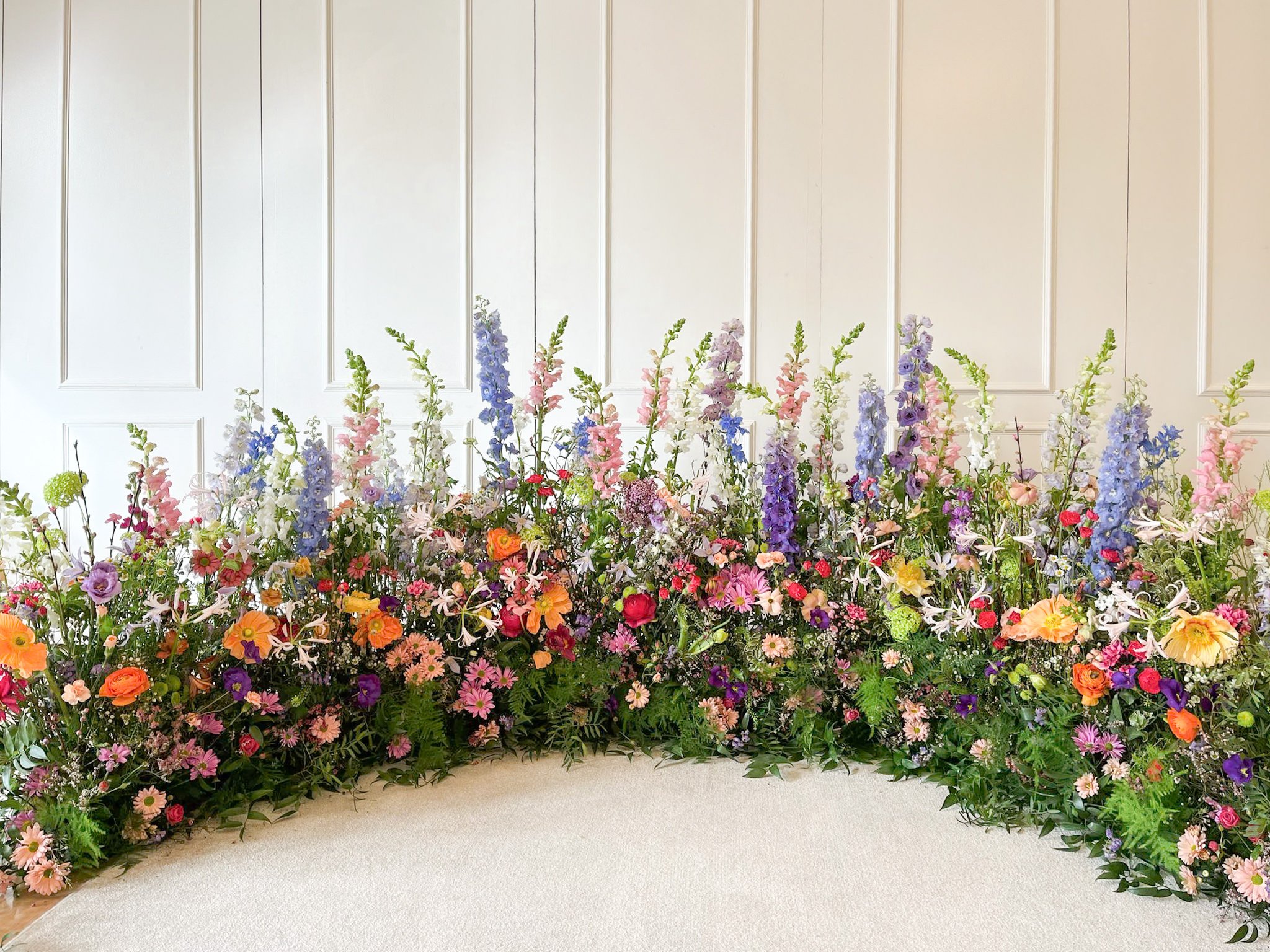 Colorful flower arrangement with mixed pink, purple, orange, and white flowers on a beige carpet in front of a white paneled wall.