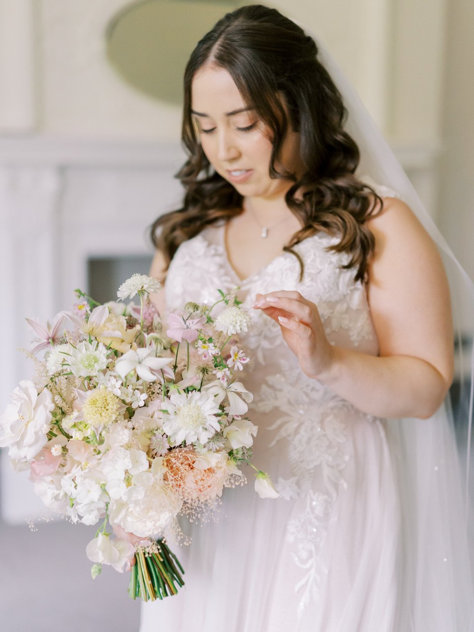 A bride in a white wedding dress holding a bouquet of white, pink, and cream flowers while adjusting a flower in her bouquet.
