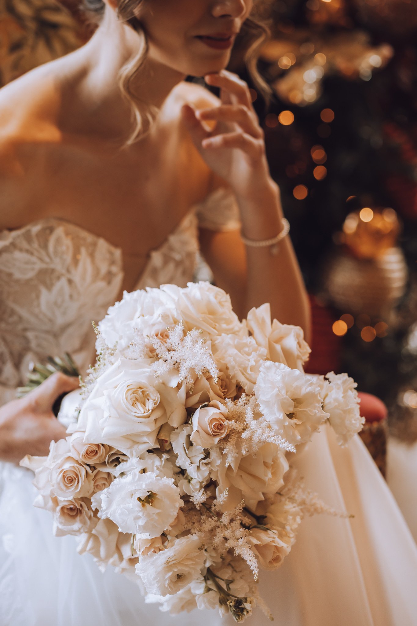A bride in a lace wedding dress holding a bouquet of white and blush roses and flowers, with her face partially visible and a Christmas tree in the background.