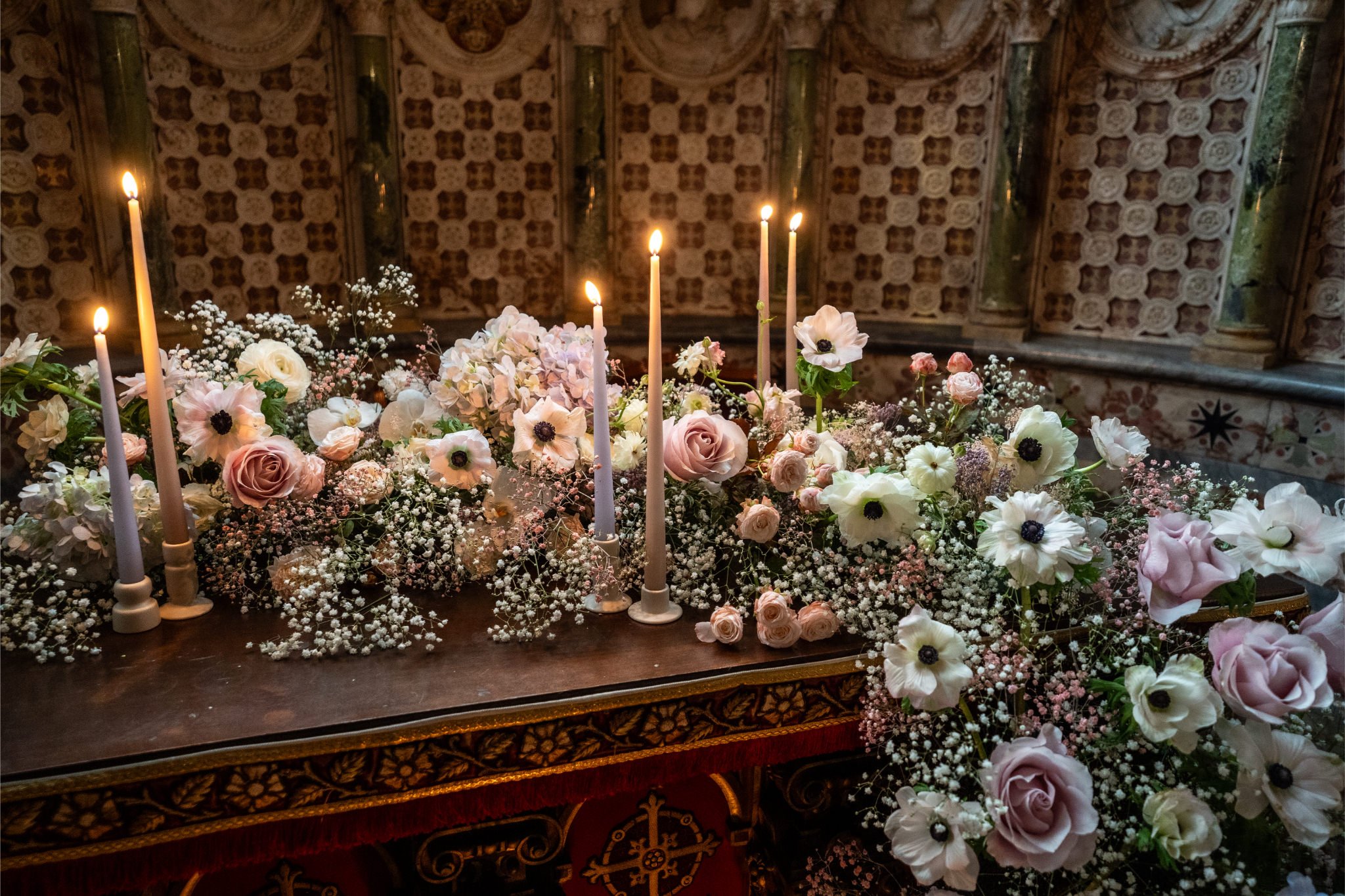 A floral arrangement of pale pink roses, white anemones, baby's breath, and eucalyptus leaves on a wooden altar with lit taper candles in a church with ornate wall designs.