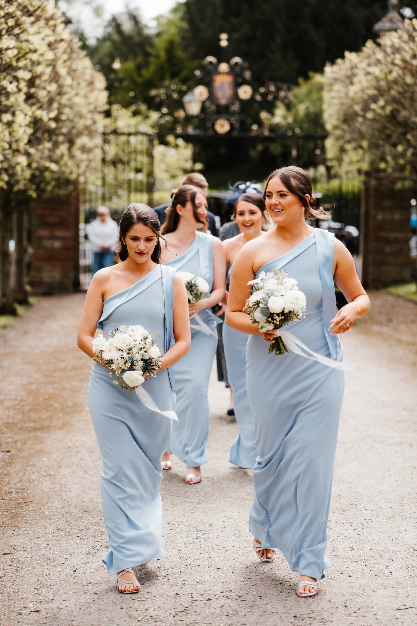 A group of women in matching light blue dresses walking together with bouquets, outdoors near a gated entrance with trees and an ornamental gate in the background.