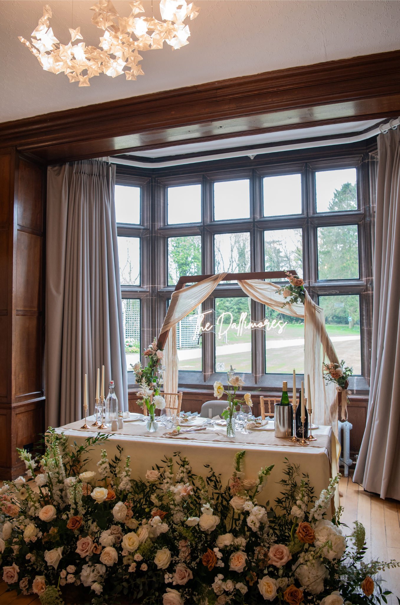 Wedding reception table with floral arrangements, candles, and a glowing neon sign reading 'The Daffiness' in front of a bay window with curtains.