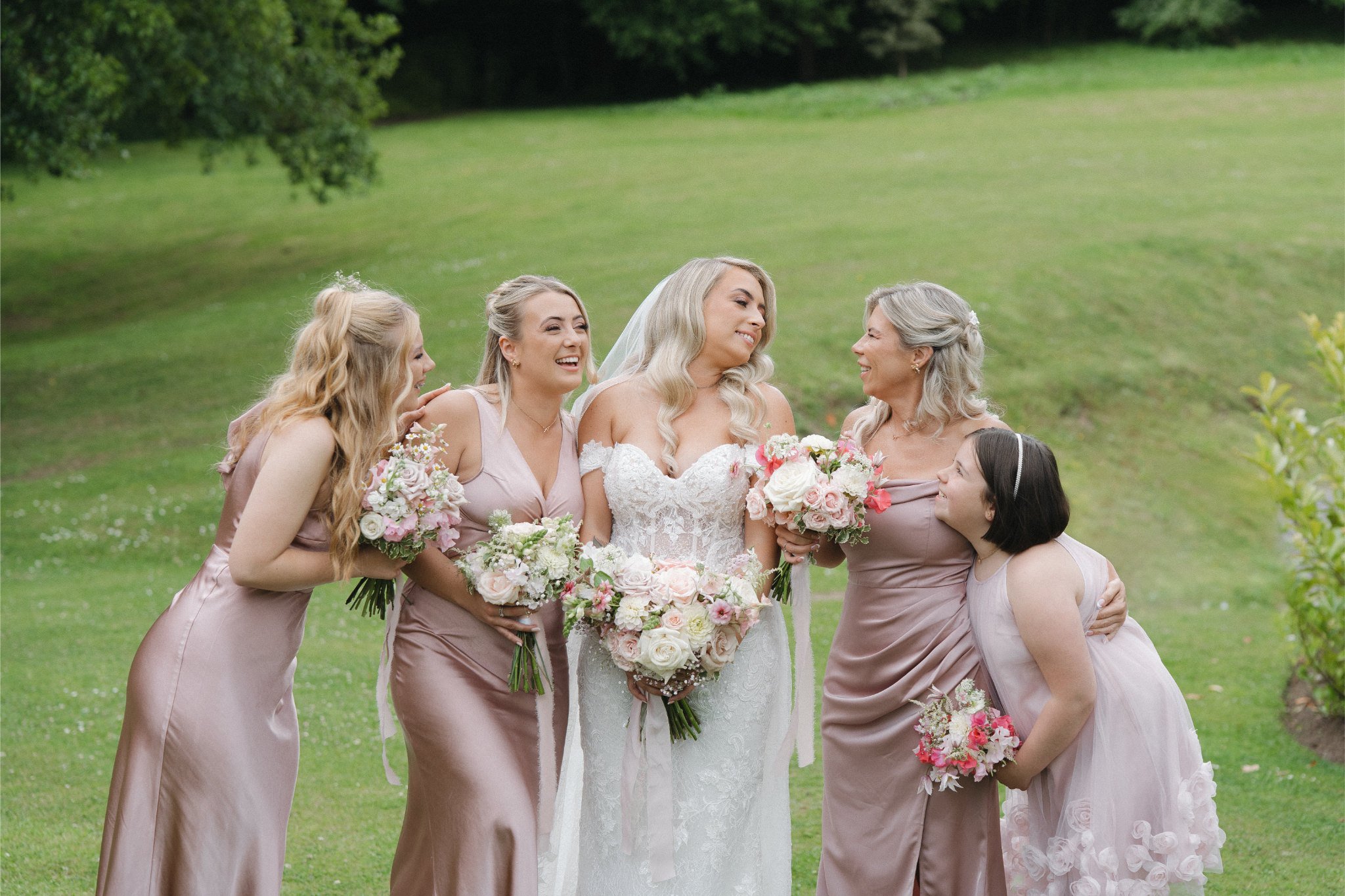 A bride with long blonde hair and her four bridesmaids, all smiling and holding bouquets, standing on a grassy lawn outdoors.
