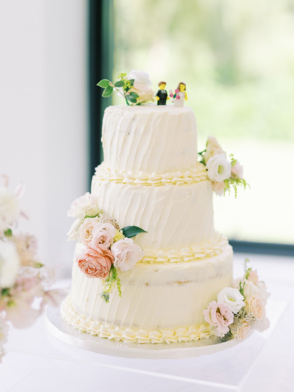 Three-tier white wedding cake decorated with light pink and white flowers, topped with a figurine of a bride and groom, placed on white tablecloth near window with blurry greenery outside.