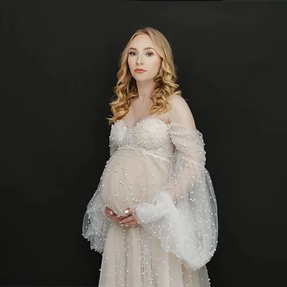 Pregnant woman with blonde hair in a flowing, off-the-shoulder, pearl-embellished dress standing against a black background