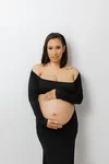 A woman with long dark hair wearing a black, off-the-shoulder dress, holding her pregnant belly.