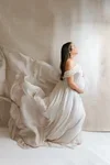 Woman sitting on a bed in a white dress, looking out a window in a softly lit room.