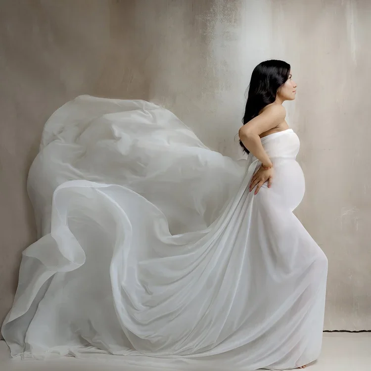 A pregnant woman in a white strapless gown standing sideways, with her hands on her lower back, against a neutral wall background.