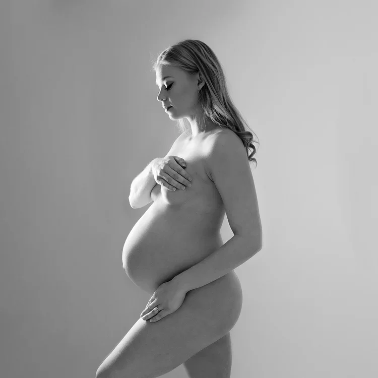 A pregnant woman standing sideways, topless, with one hand covering her chest and the other resting on her belly, in a black and white photo.