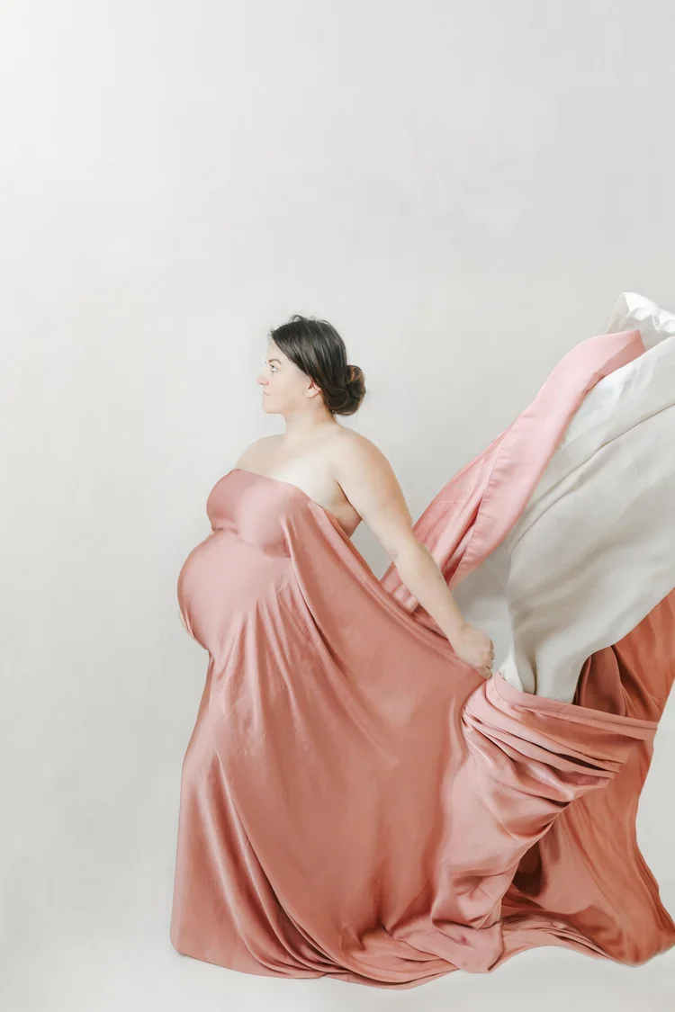 Pregnant woman dressed in pink satin fabric, sitting with her back to a bed, holding the fabric with her hands.