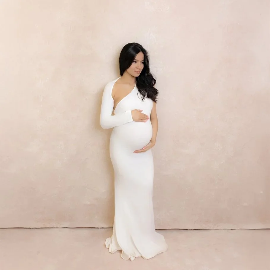 A pregnant woman with long dark hair wearing a white gown, standing against a light beige background, gently holding her belly.