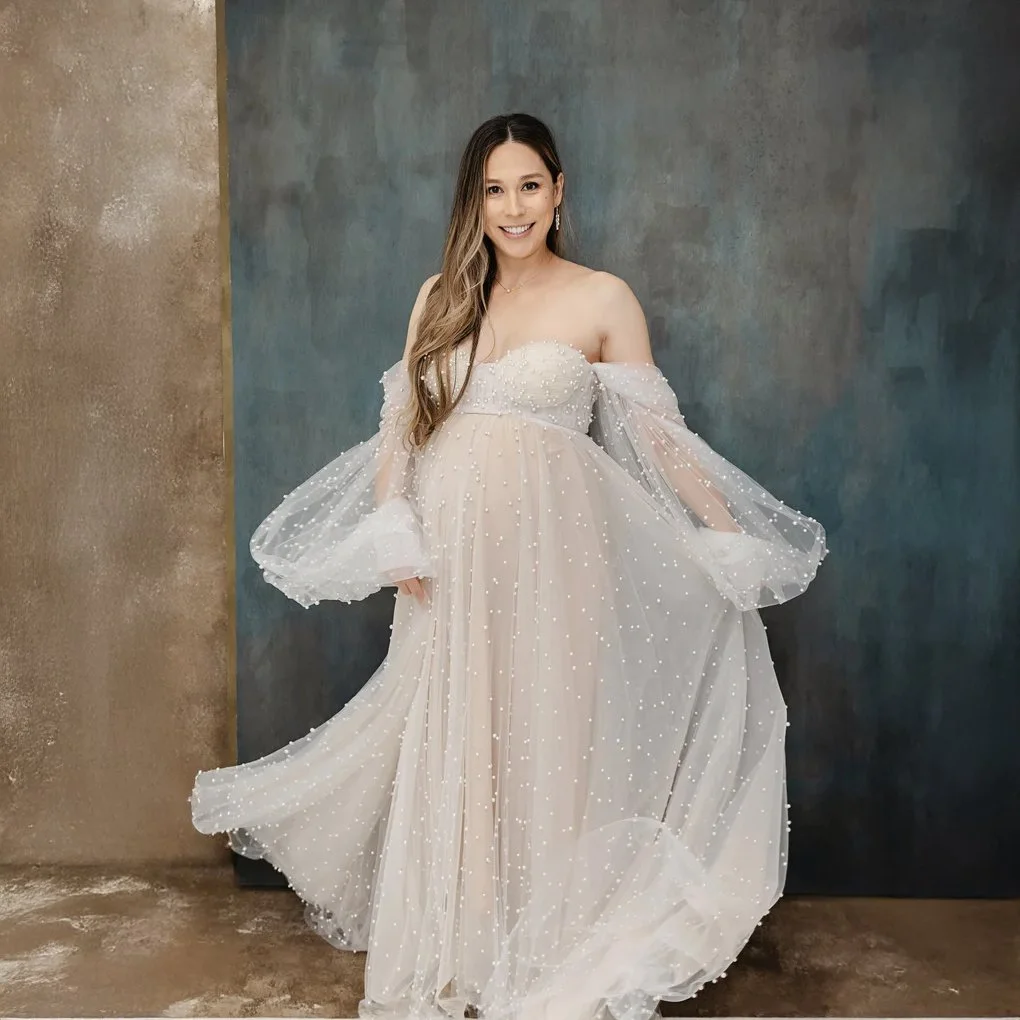 A woman in a white, off-the-shoulder, pearl-adorned wedding dress standing against a dark blue wall, smiling and holding the dress.