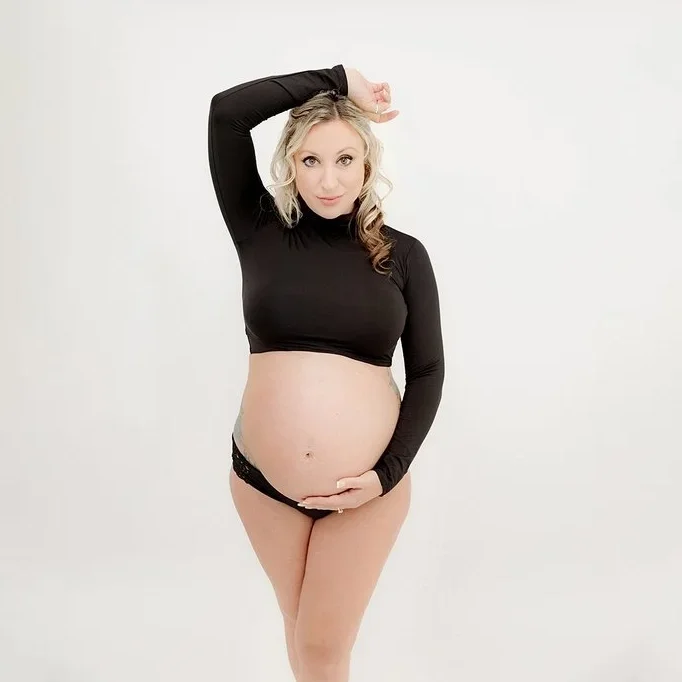 Pregnant woman in black long-sleeve crop top and underwear posing against a plain white background.