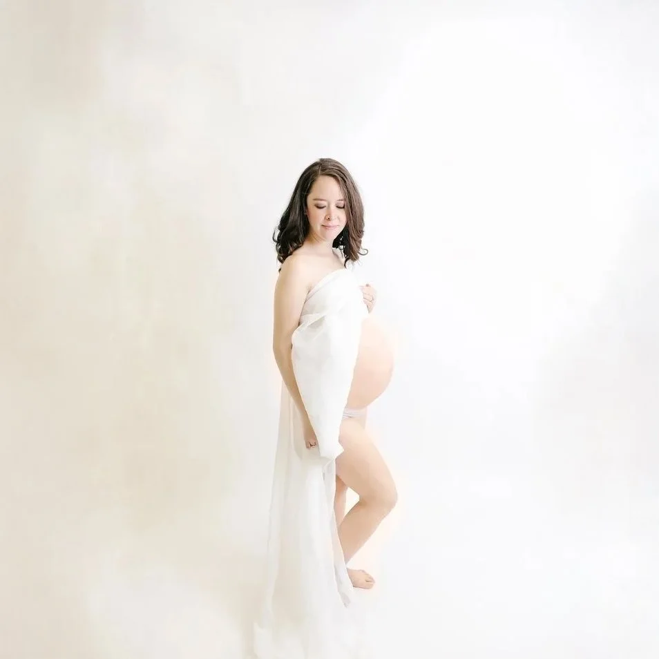 Pregnant woman wrapped in white fabric standing against a plain white background.