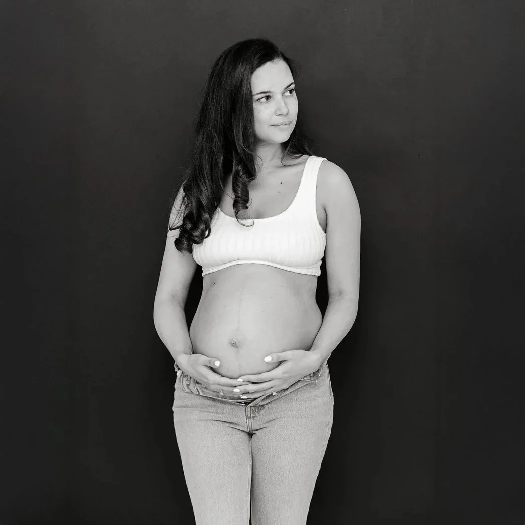 Black and white photo of a pregnant woman wearing a white tank top and jeans, standing against a dark background, with her hands gently resting on her pregnant belly.