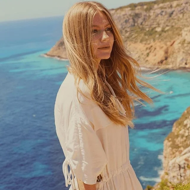 Woman in white dress standing on a cliff overlooking the ocean with clear blue water and rocky coastline under a bright sky.