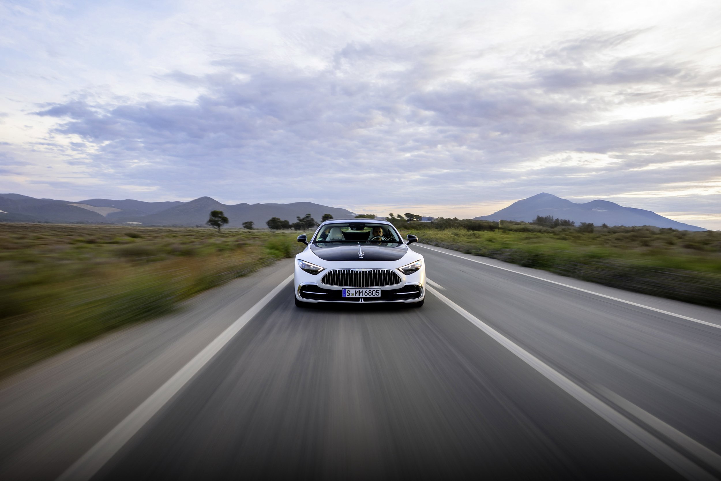A silver Mercedes-Benz Maybach driving on an open road through scenic countryside with mountains in the background.