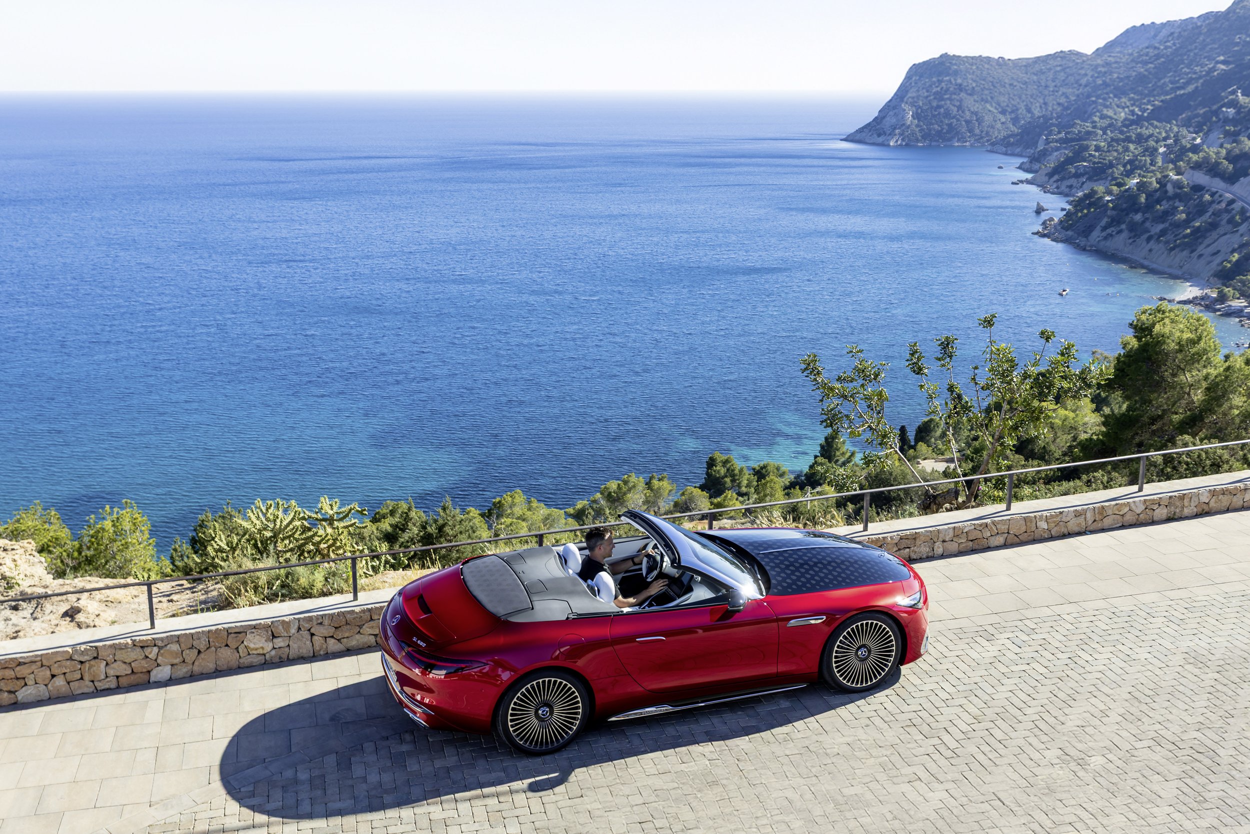 Red convertible car parked on a paved overlook with a scenic coastal view and mountain in the background.