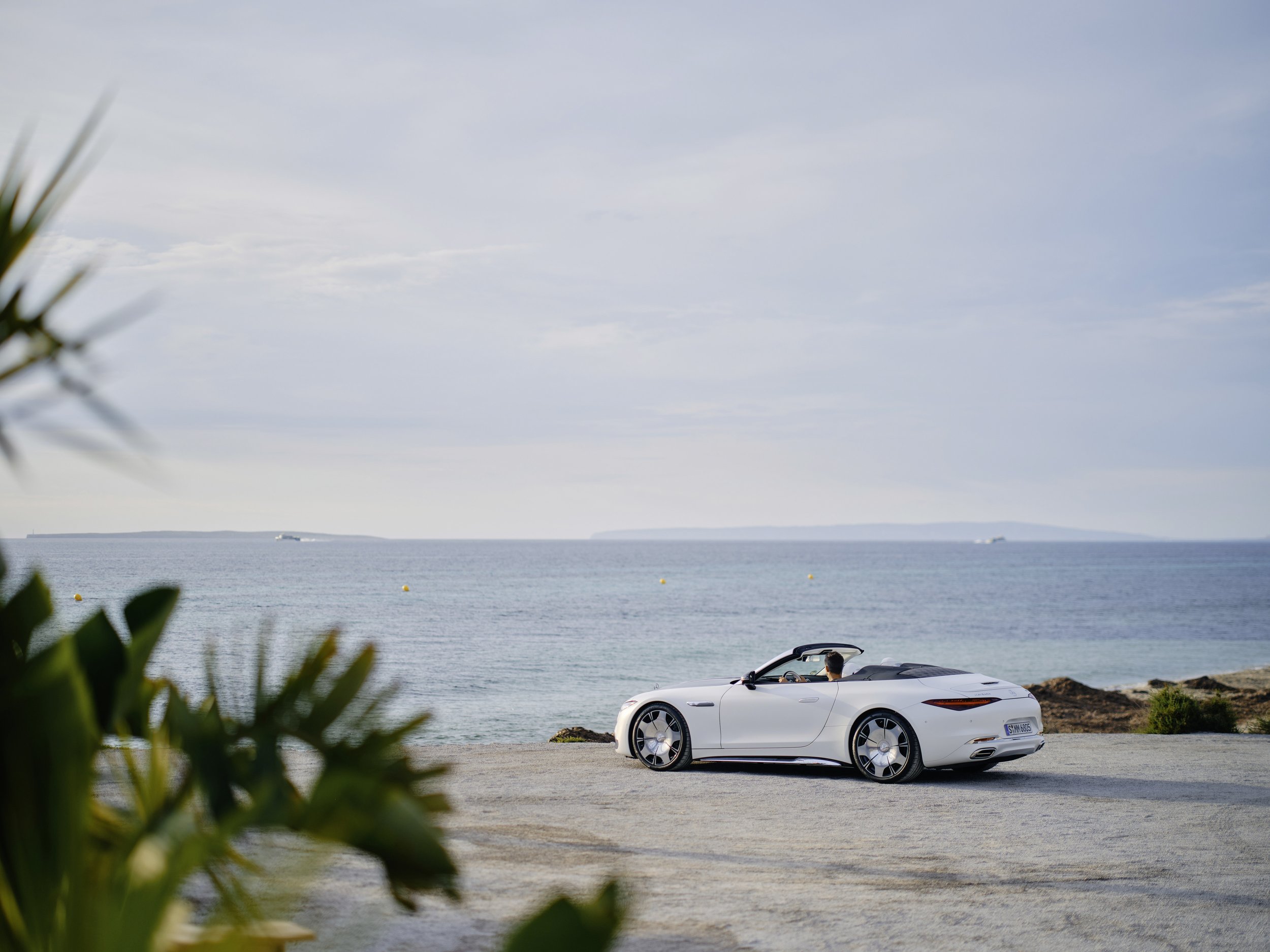 A white convertible sports car parked on a gravel area near the ocean with a distant view of land across the water and some plants in the foreground.