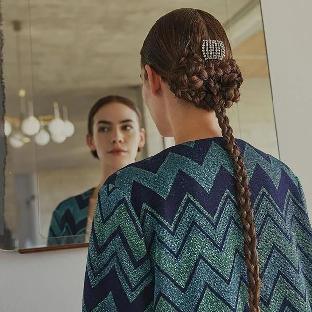 Person with long braided hair and zigzag-patterned sweater looking in a mirror.