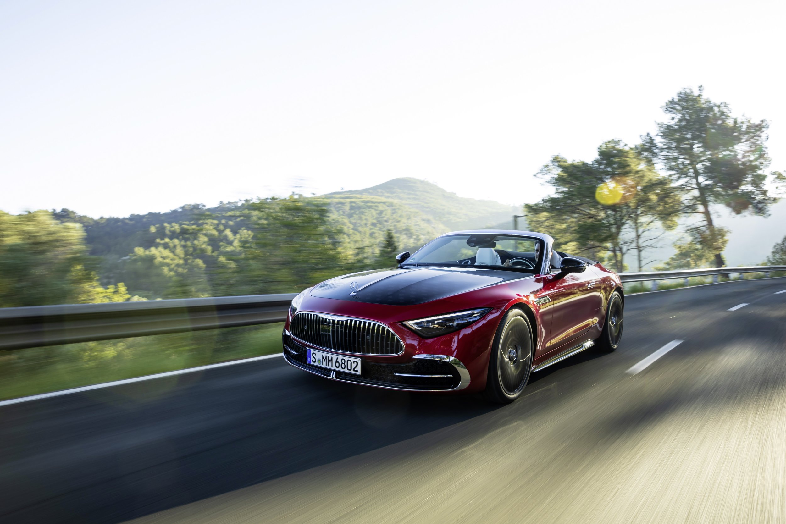 A red Mercedes-Benz convertible driving on a scenic mountain road with trees and hills in the background.