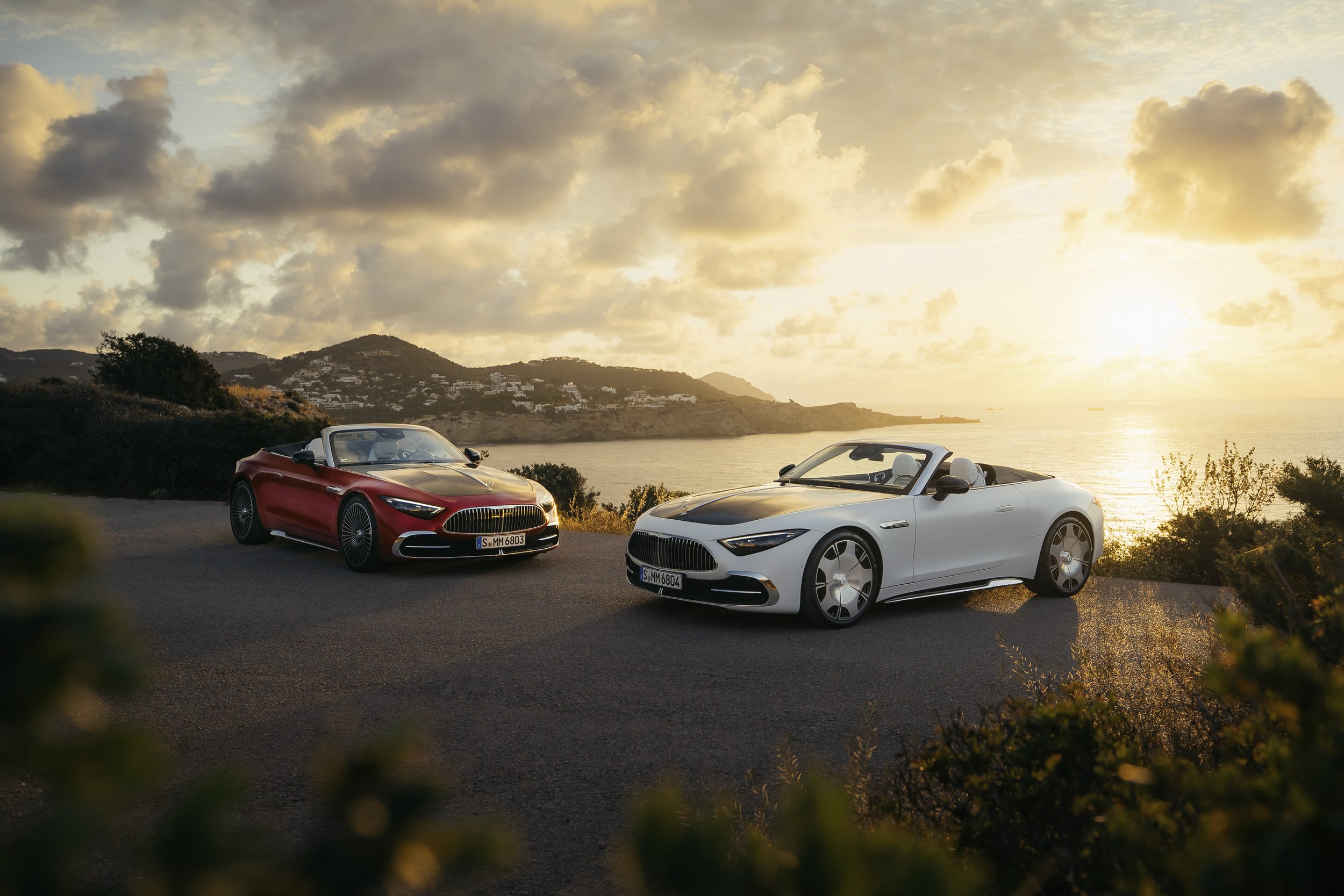 Two modern convertible cars, one red and one white, parked on a coastal road with a sunset over the ocean in the background.