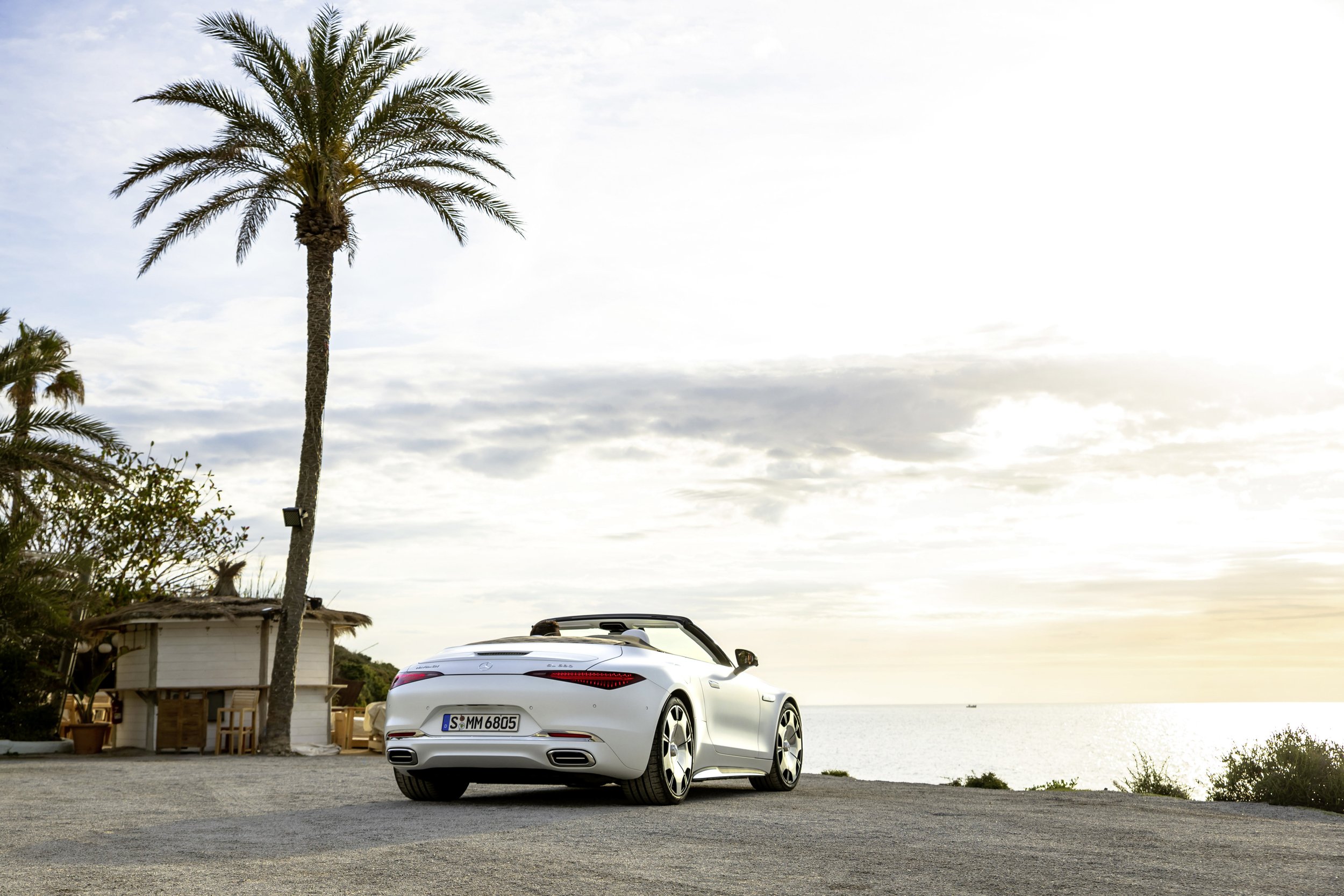 A white convertible sports car parked on a coastal area during sunset with palm trees and a small hut nearby.