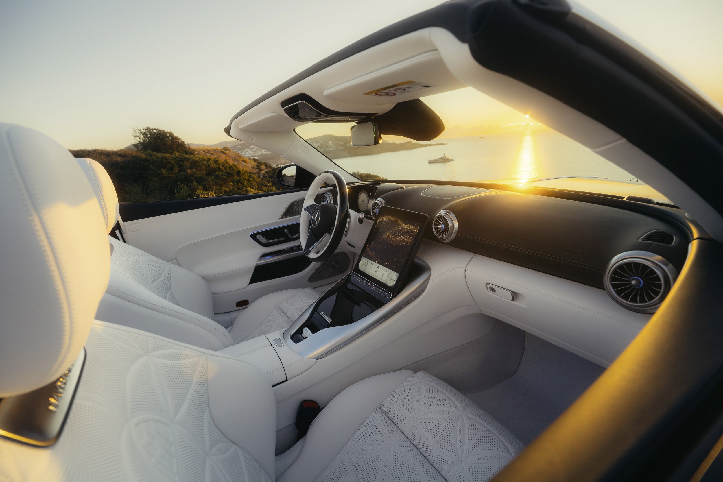 Interior of a luxury convertible car with white leather seats, dashboard, and a large touchscreen display during sunset over a coastal landscape.