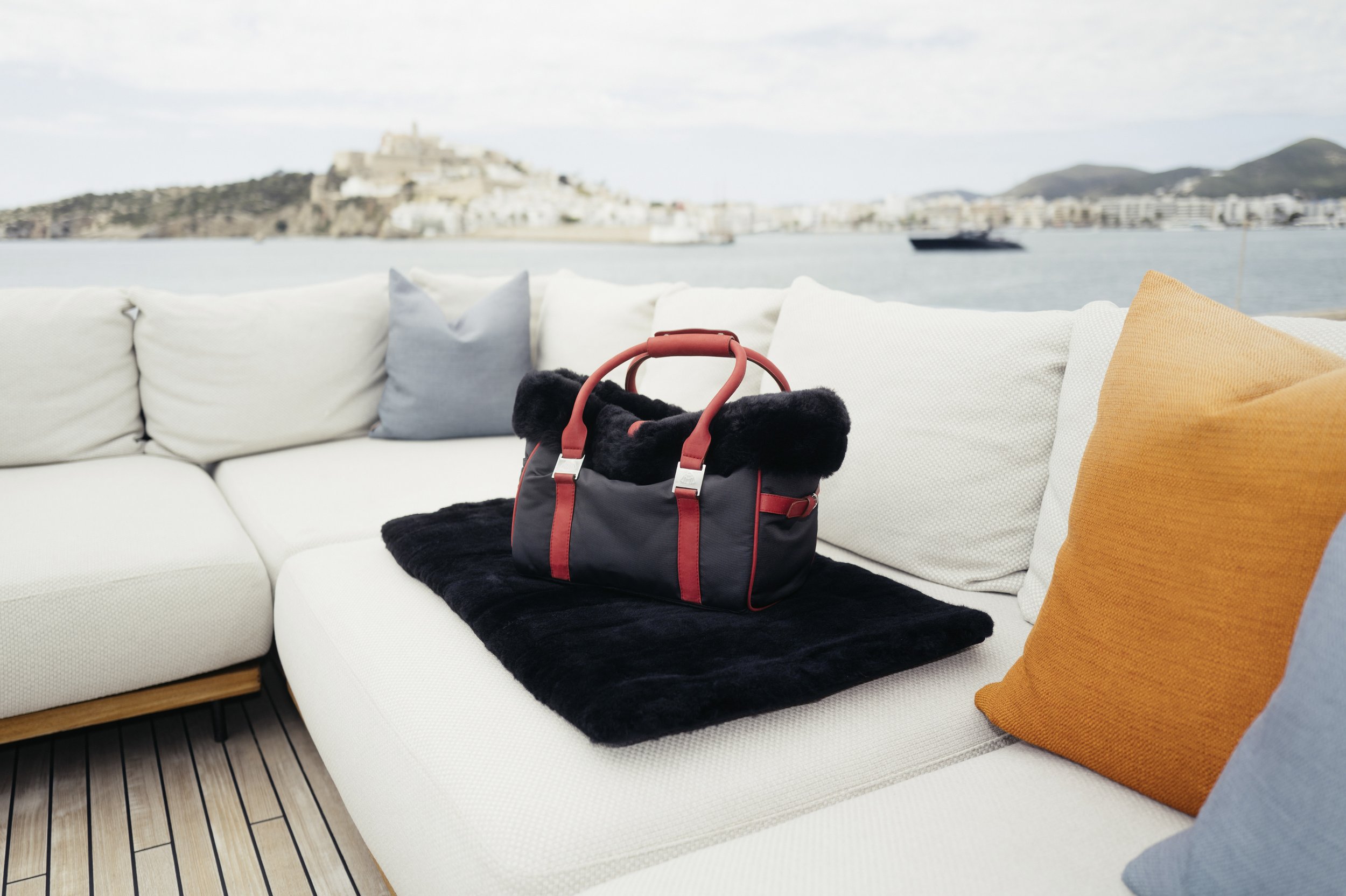 A black and red duffel bag with fur trims resting on a black blanket on a white outdoor sofa with colorful pillows, overlooking a body of water and a distant island or fortress.