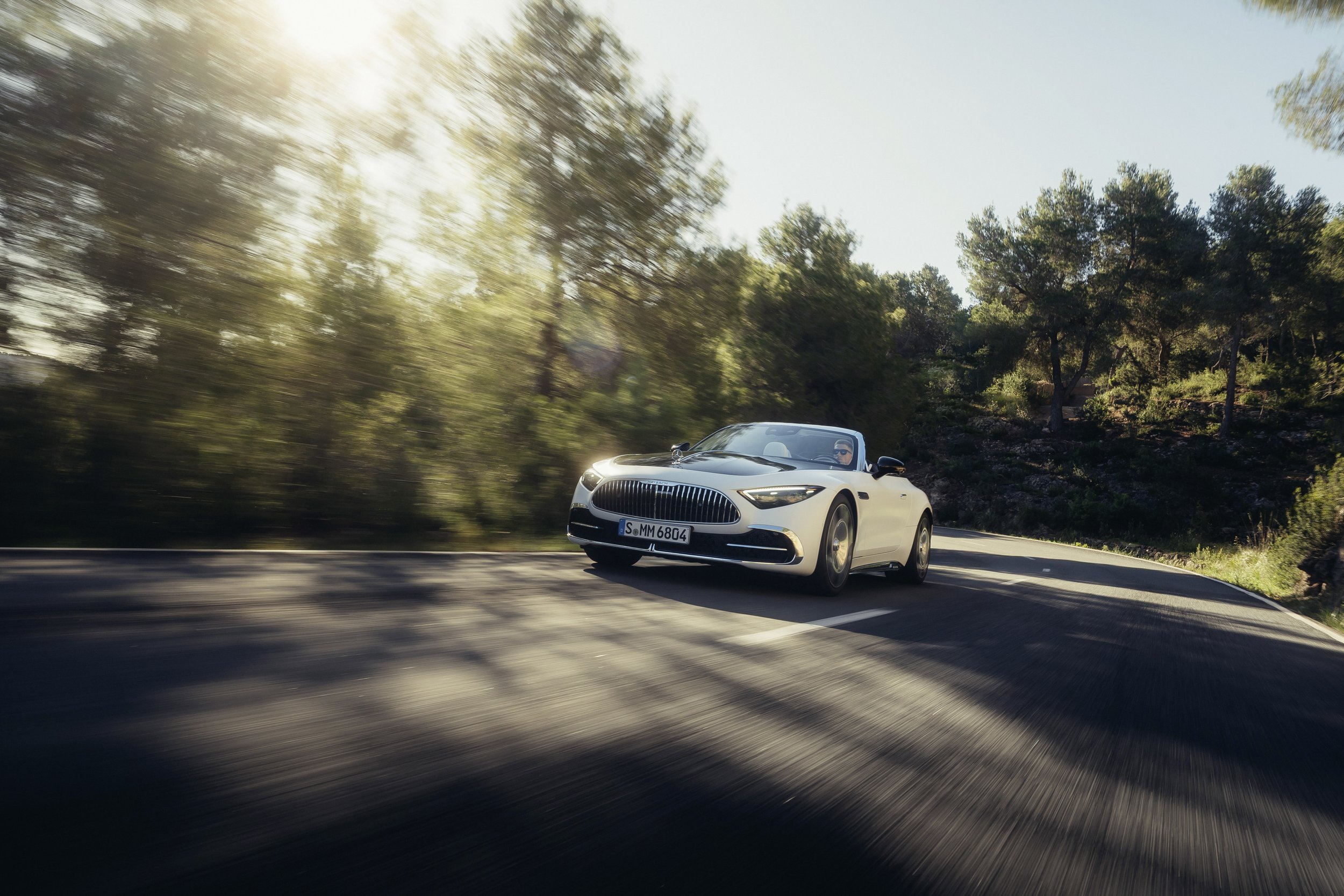 A white convertible car driving on a winding road surrounded by trees with sunlight filtering through.