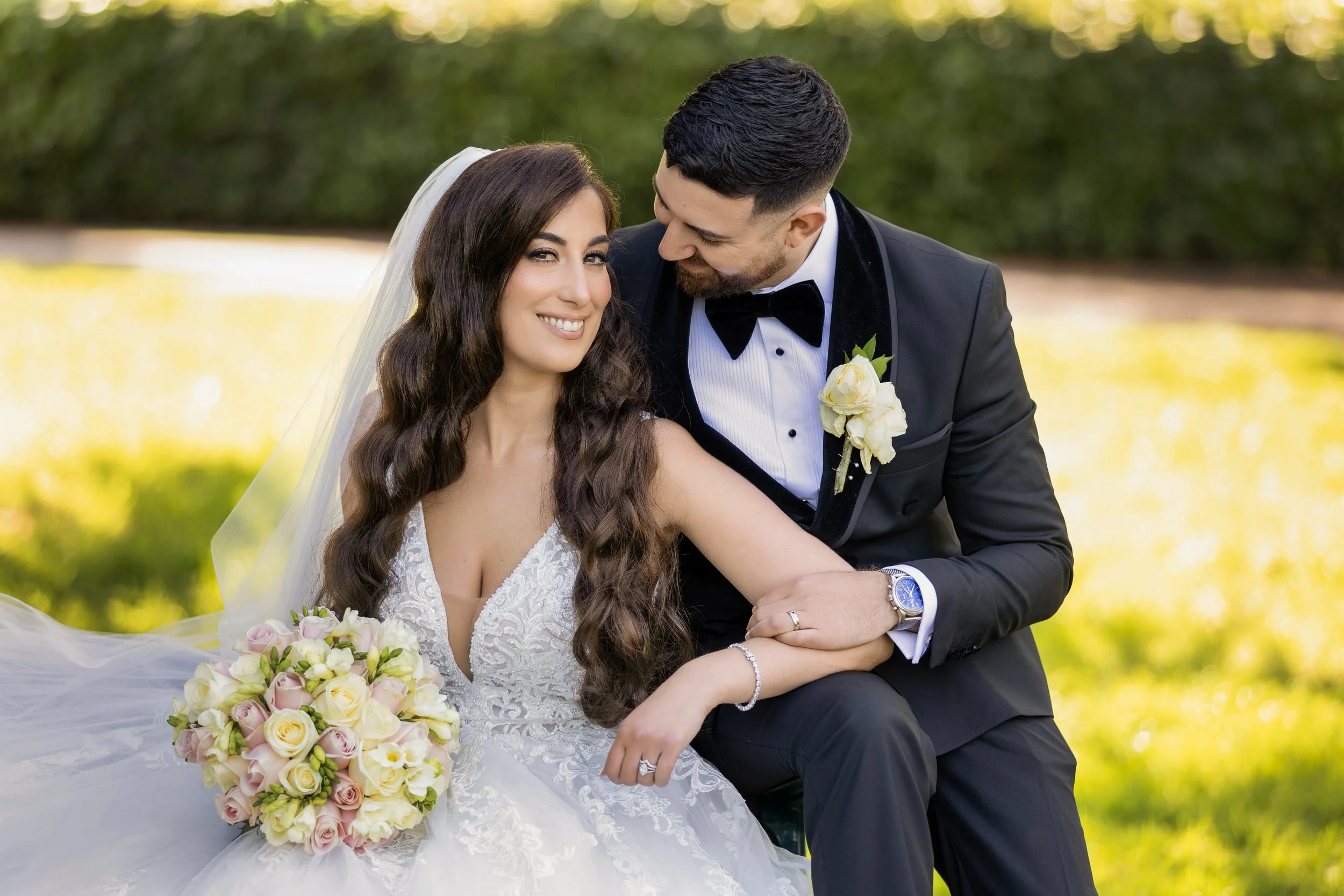A bride and groom sitting outdoors, the groom leaning in towards the bride who is smiling, holding a bouquet of pink and white roses, surrounded by greenery and sunlight.