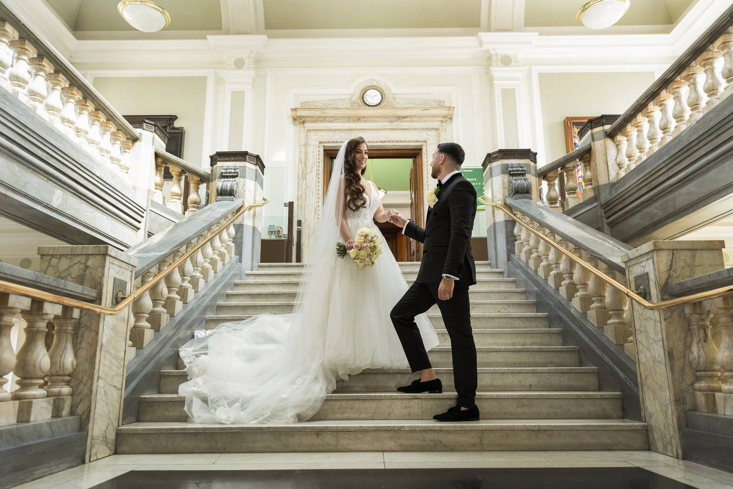 A bride and groom standing on a marble staircase inside a grand building, holding hands and looking at each other. The bride is wearing a white wedding gown and veil, holding a bouquet of pink and white roses. The groom is dressed in a black tuxedo with a white shirt and black bow tie.