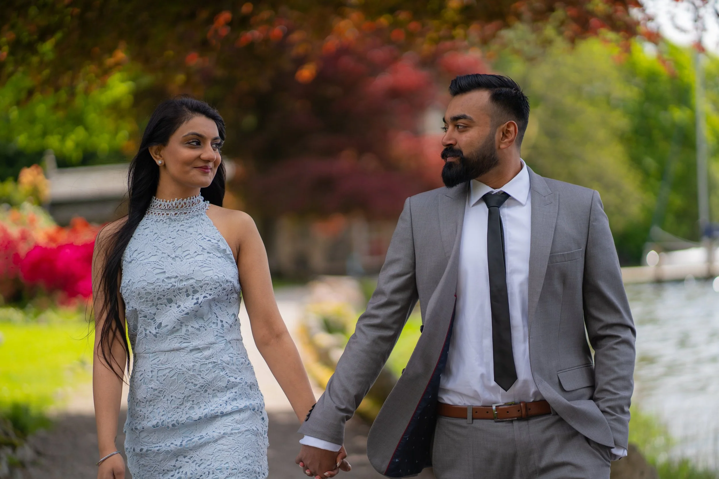 A man and woman holding hands and walking outdoors in a park, with colorful autumn trees in the background.