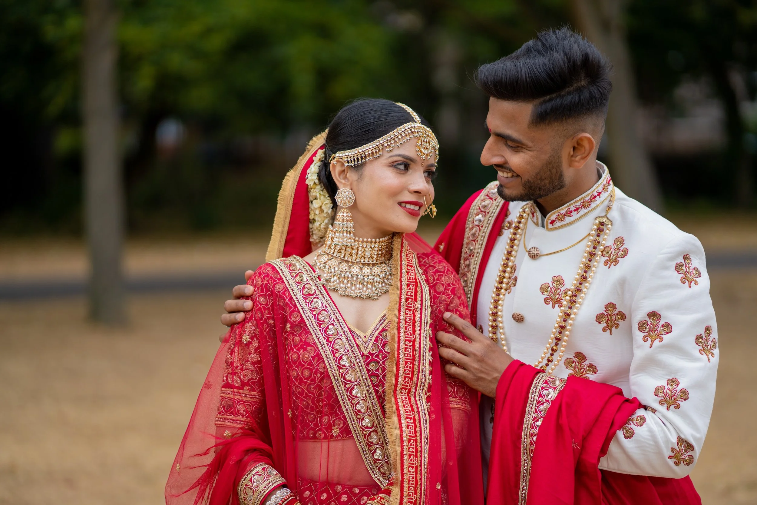 A couple dressed in traditional Indian wedding attire, smiling and gazing at each other outdoors with a blurred green background.