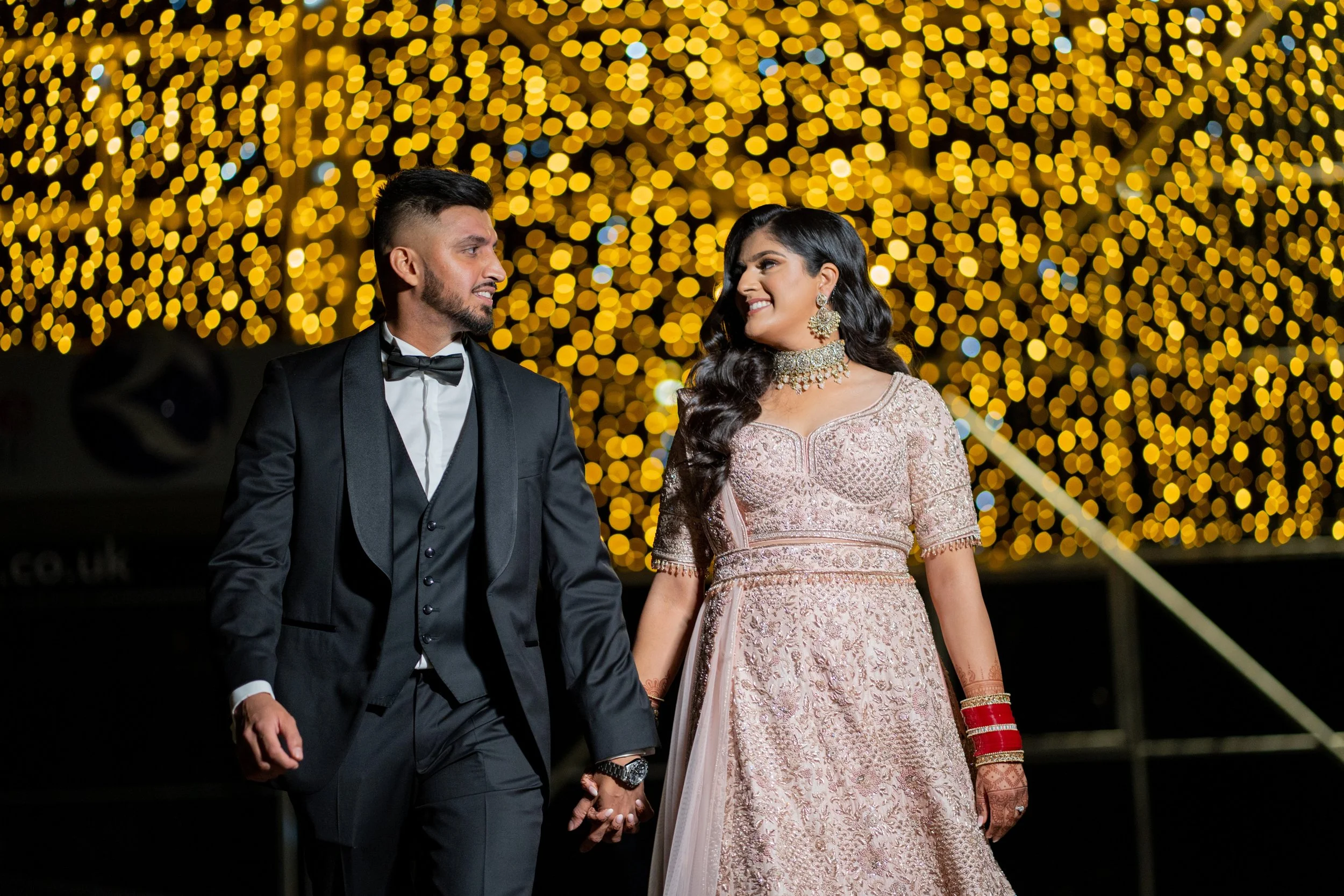 A couple dressed in formal attire holding hands at a celebration, with a background of blurred golden string lights.