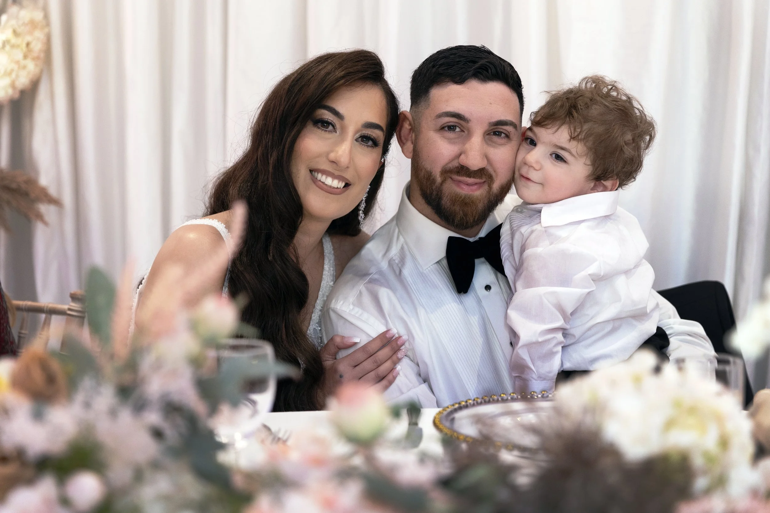 A happy family of three, including a woman with long brown hair, a man with short black hair and a beard, and a young boy with curly brown hair, dressed in formal attire at a celebration or wedding, sitting at a decorated table with blurred flowers in the foreground.