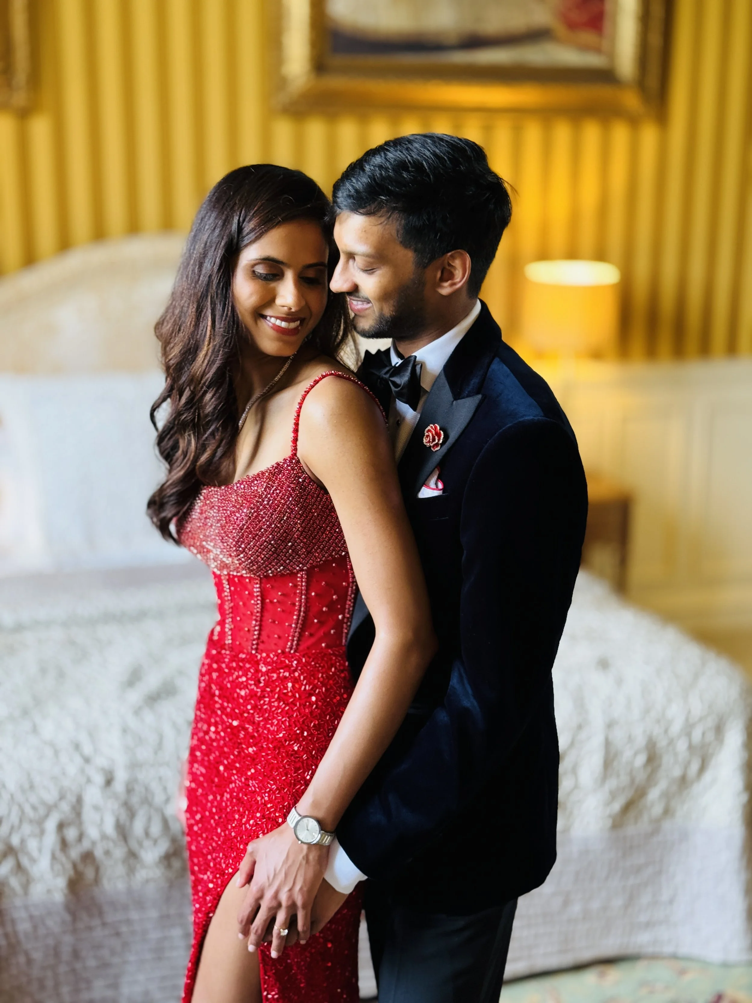 A woman in a red, sequined gown and a man in a tuxedo pose together in an elegant room with gold and beige decor.