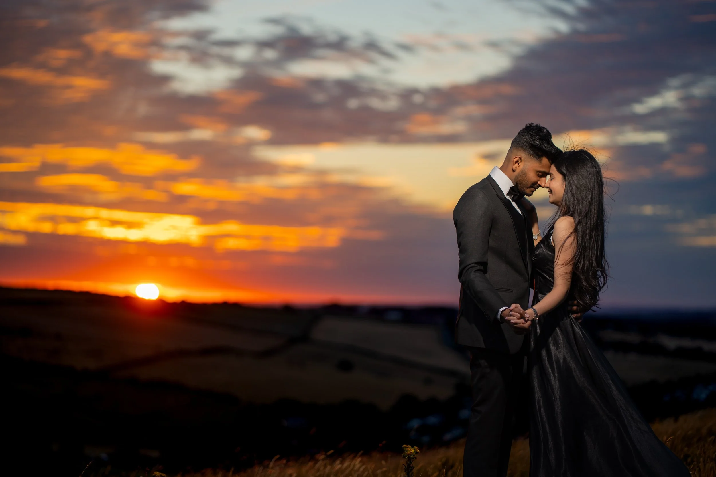 A man and woman in formal attire holding hands and touching foreheads, smiling affectionately against a sunset sky with colorful clouds and a glowing horizon.