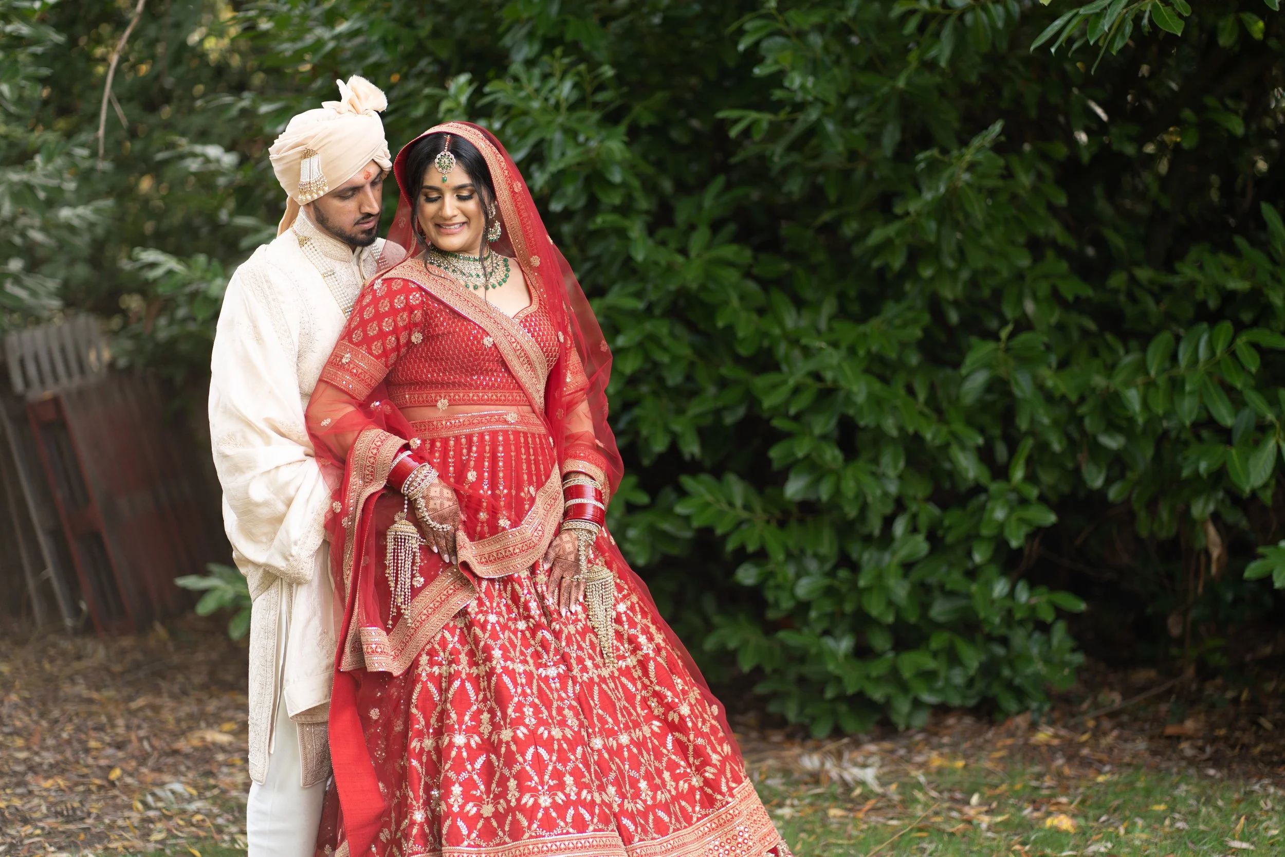 An Indian couple in traditional wedding attire standing outdoors near green bushes. The bride wears a red lehenga with gold embroidery and matching jewelry. The groom wears a cream-colored sherwani and turban.