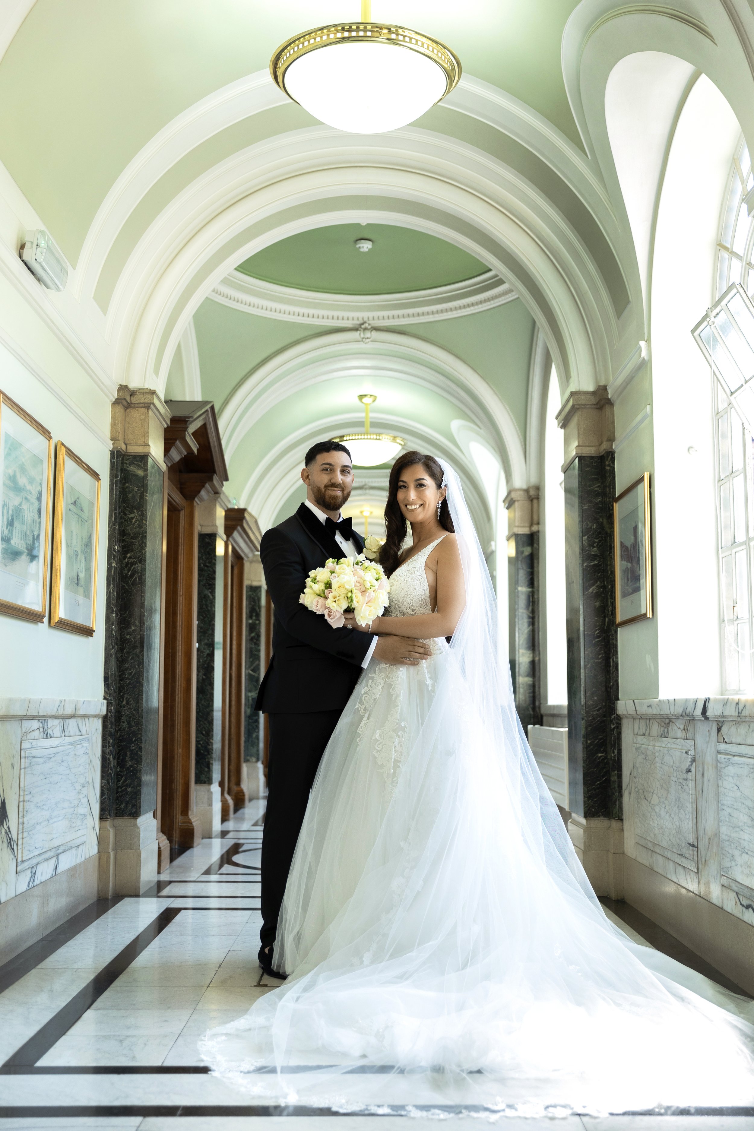 A newlywed couple in wedding attire standing in a beautifully lit hallway with arched ceilings, marble floors, and framed pictures on the walls, sharing a joyful moment.