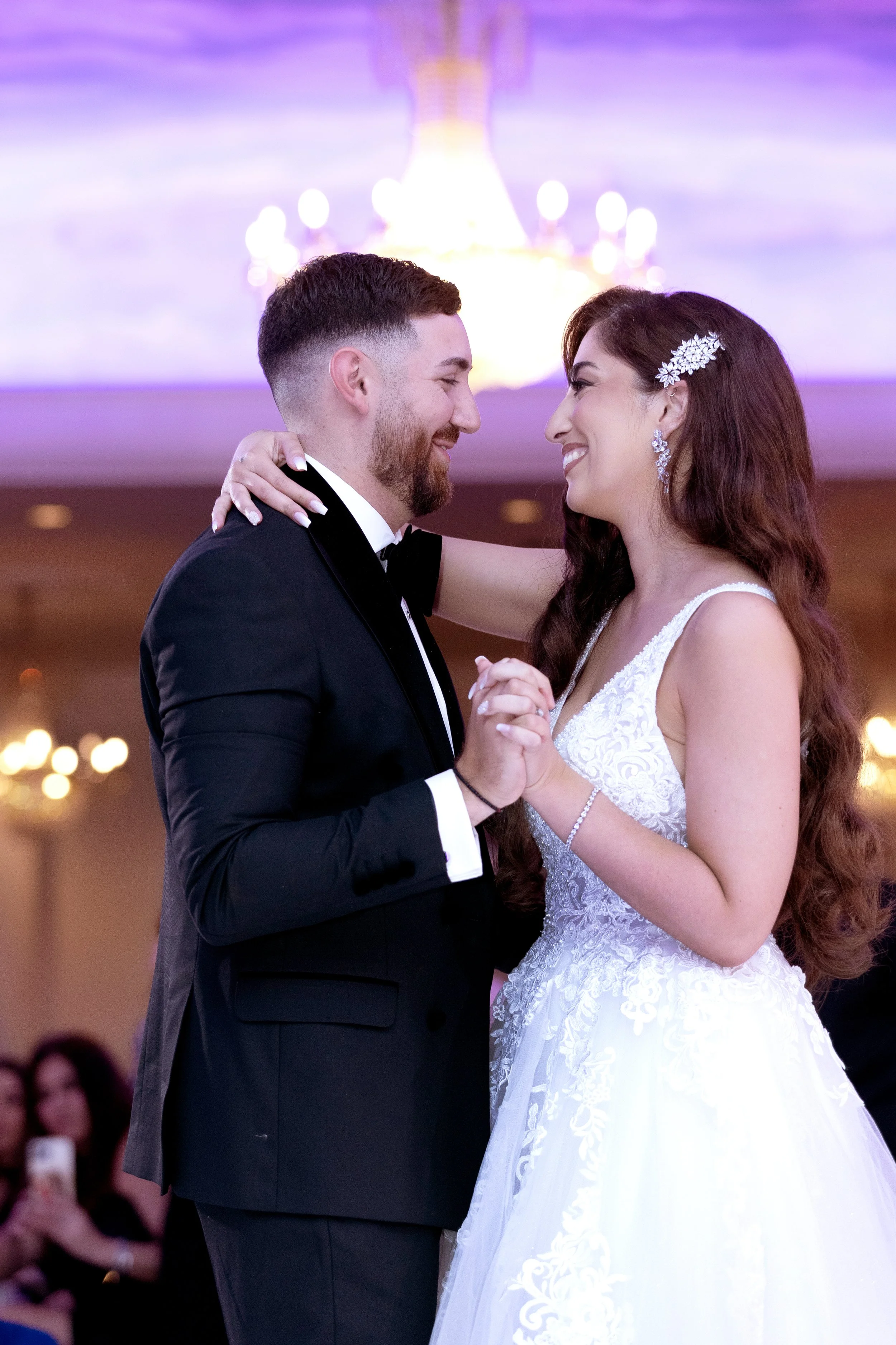 A bride and groom share a dance at their wedding reception. The bride has long wavy hair with a floral hairpiece and wears a white lace wedding gown. The groom has short dark hair and a beard, and is dressed in a black tuxedo with a bow tie. They are smiling and looking into each other's eyes, holding hands and embracing in a warmly lit venue with a chandelier in the background.