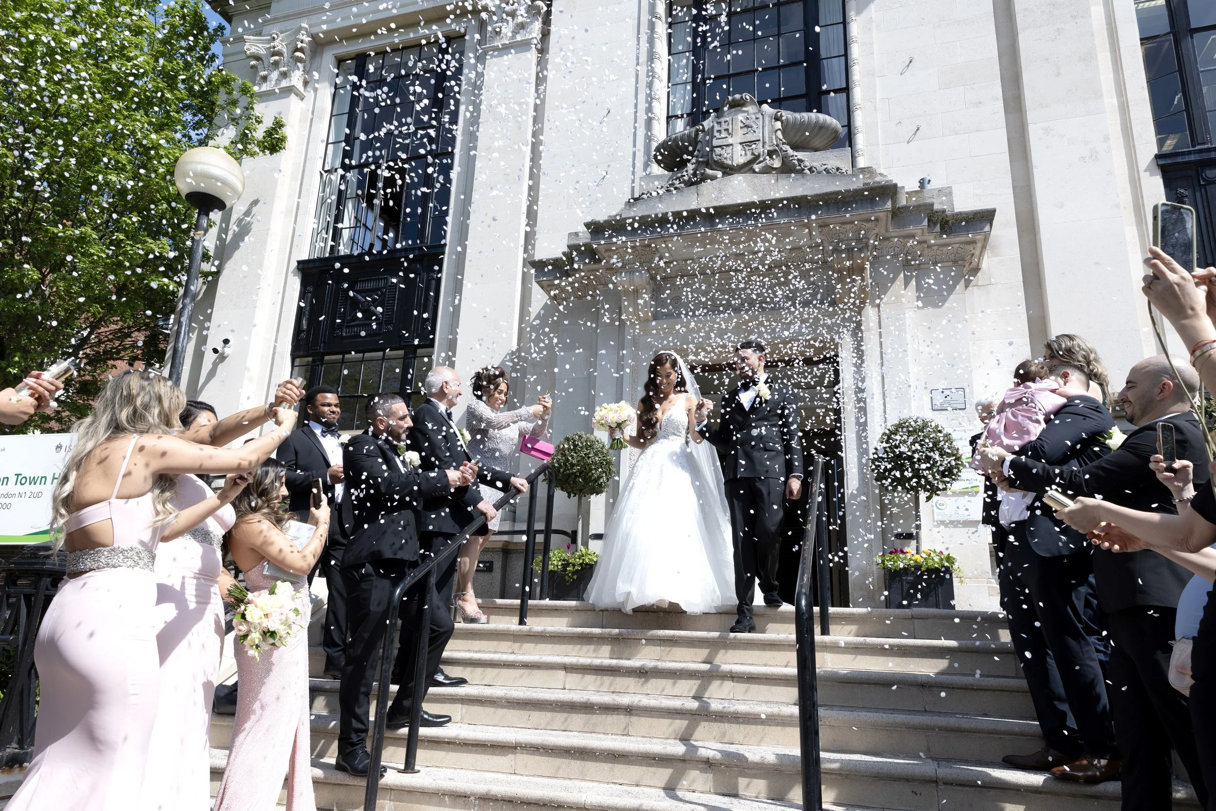 A wedding celebration outside a white stone building with classical architecture features the bride and groom standing on steps among guests. Guests are throwing confetti, and some are holding phones and cameras. The bride wears a white wedding dress, and the groom wears a tuxedo. The scene takes place on a sunny day with green trees nearby.