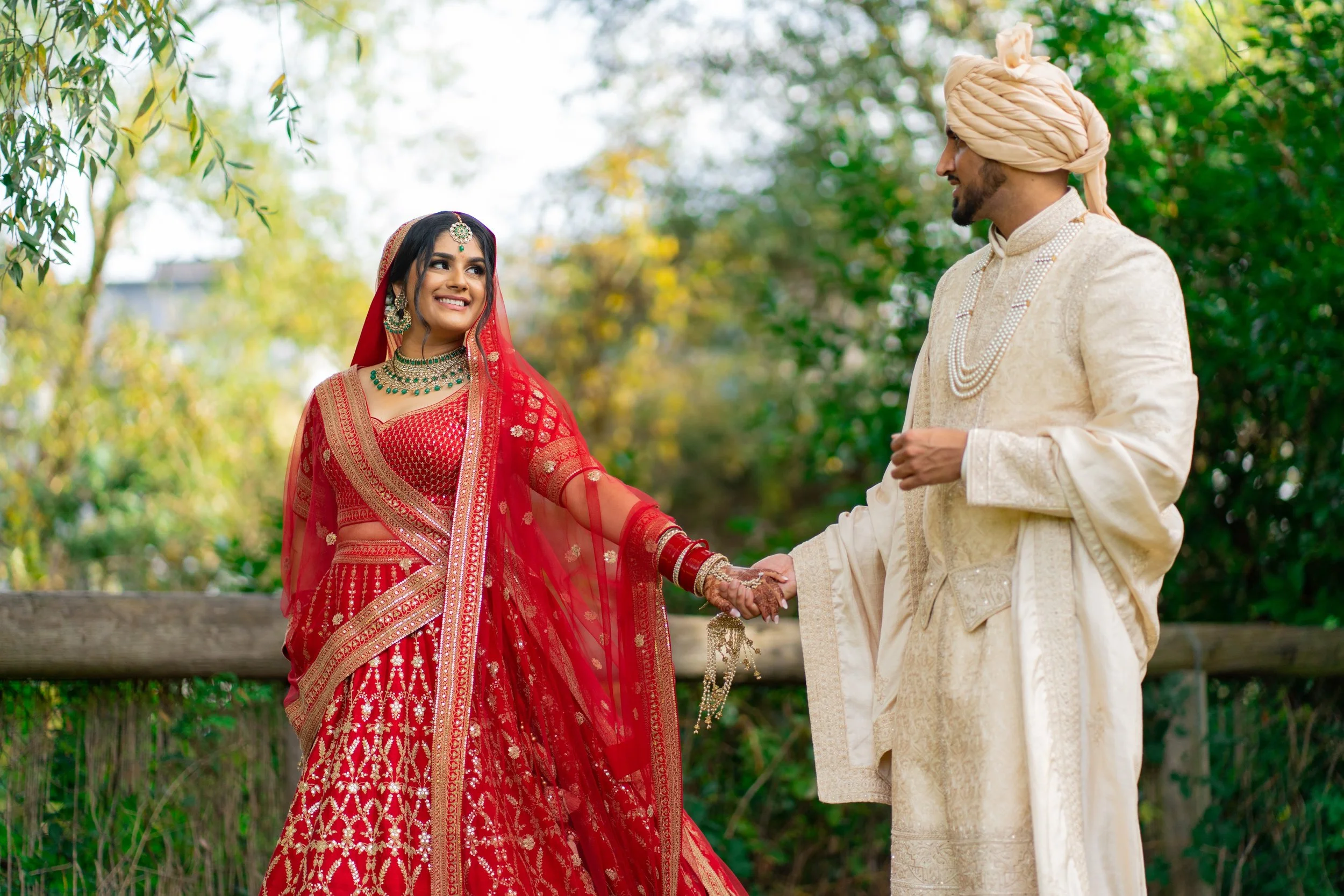 A wedding couple outdoors with the bride in a red traditional dress and jewelry, and the groom in a cream traditional outfit with a turban, holding hands and smiling at each other.
