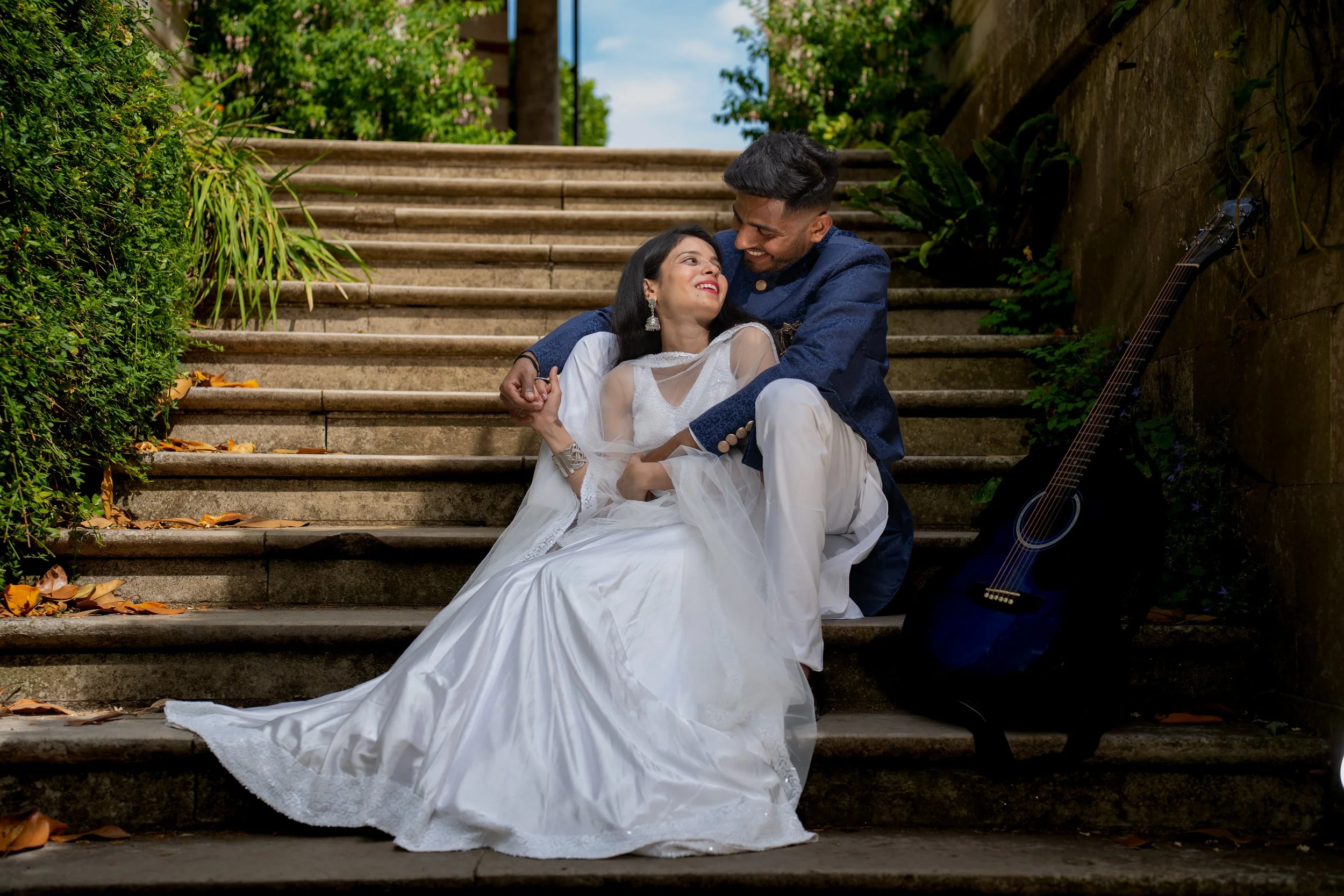 A couple dressed in traditional wedding attire sitting on outdoor stone stairs, smiling at each other with a guitar nearby and greenery around.