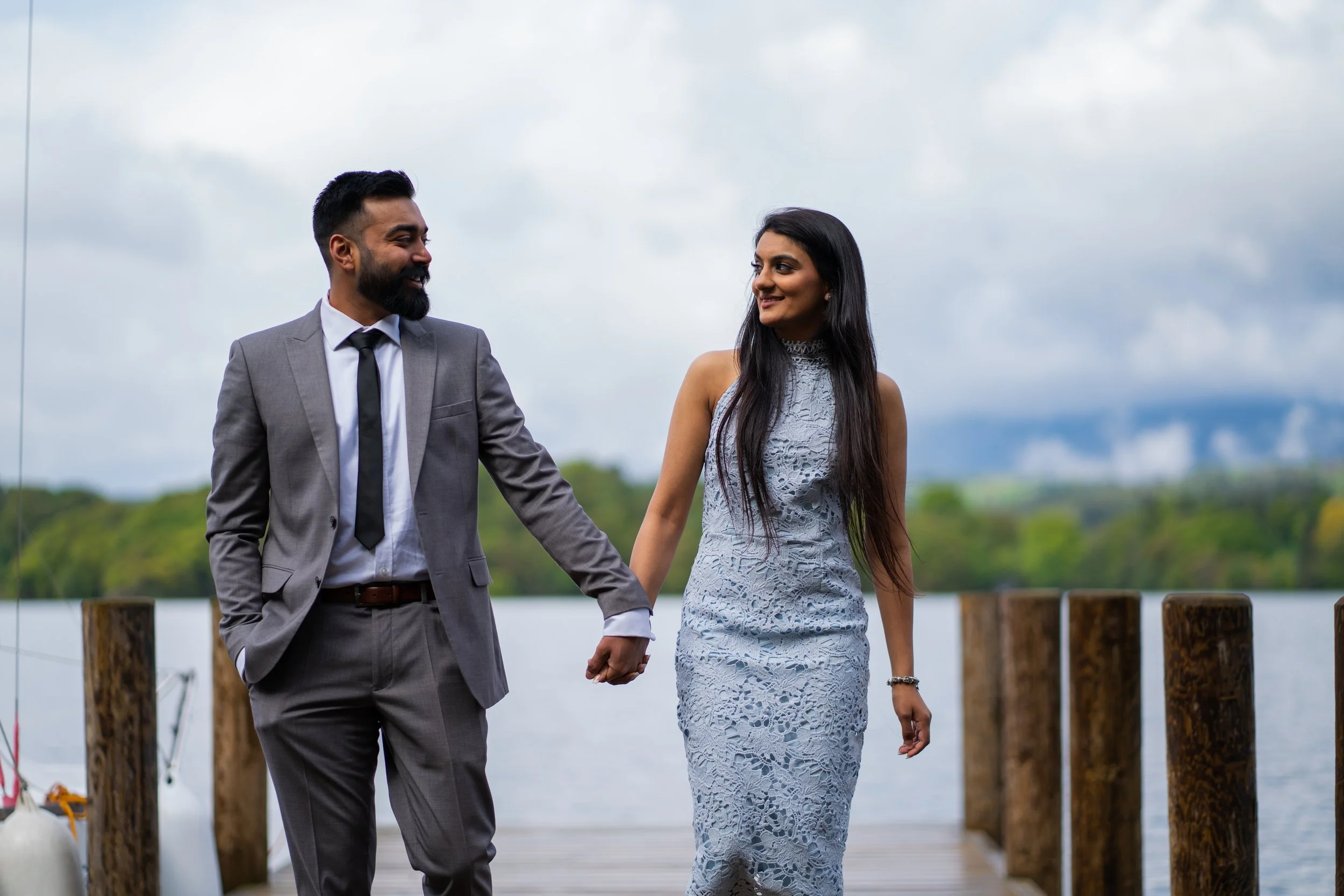 A man in a gray suit and a woman in a lace dress holding hands and smiling at each other outdoors by a lake with cloudy sky.