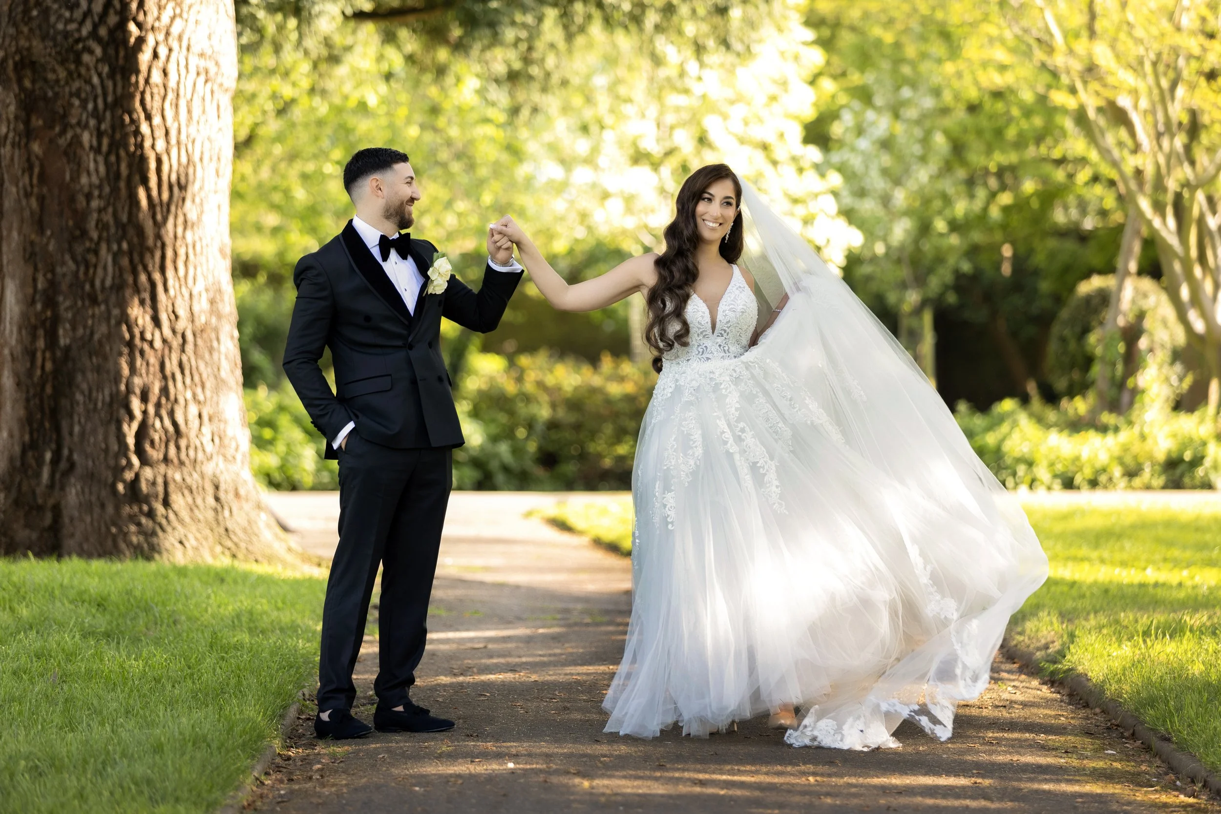 A bride and groom on their wedding day in a park, holding hands and smiling at each other. The groom wears a black tuxedo with a bow tie, and the bride wears a white wedding gown with a veil. They are standing on a pathway surrounded by green grass and trees with sunlight filtering through the leaves.