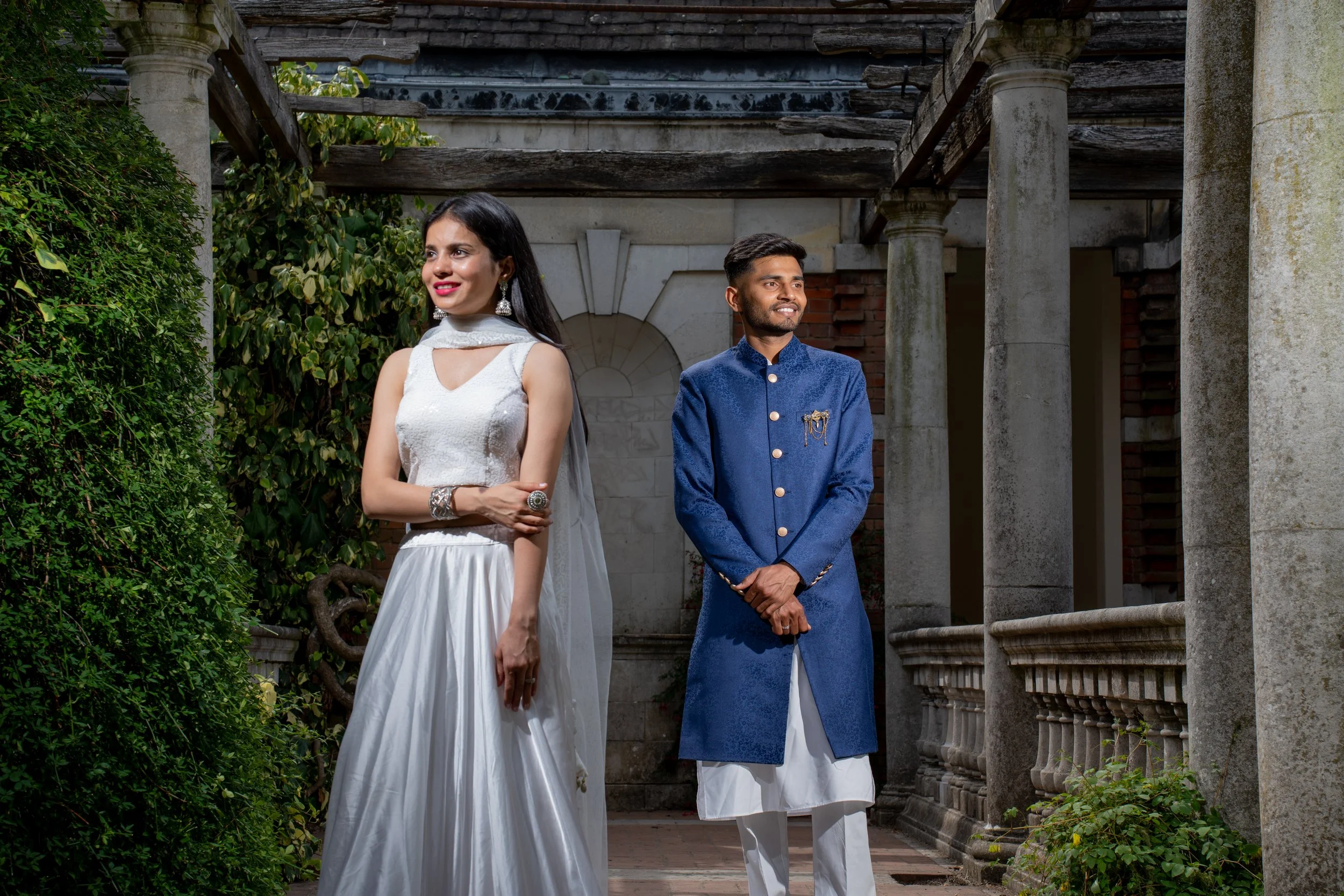 A couple dressed in traditional Indian attire standing outdoors near historical stone columns and greenery.